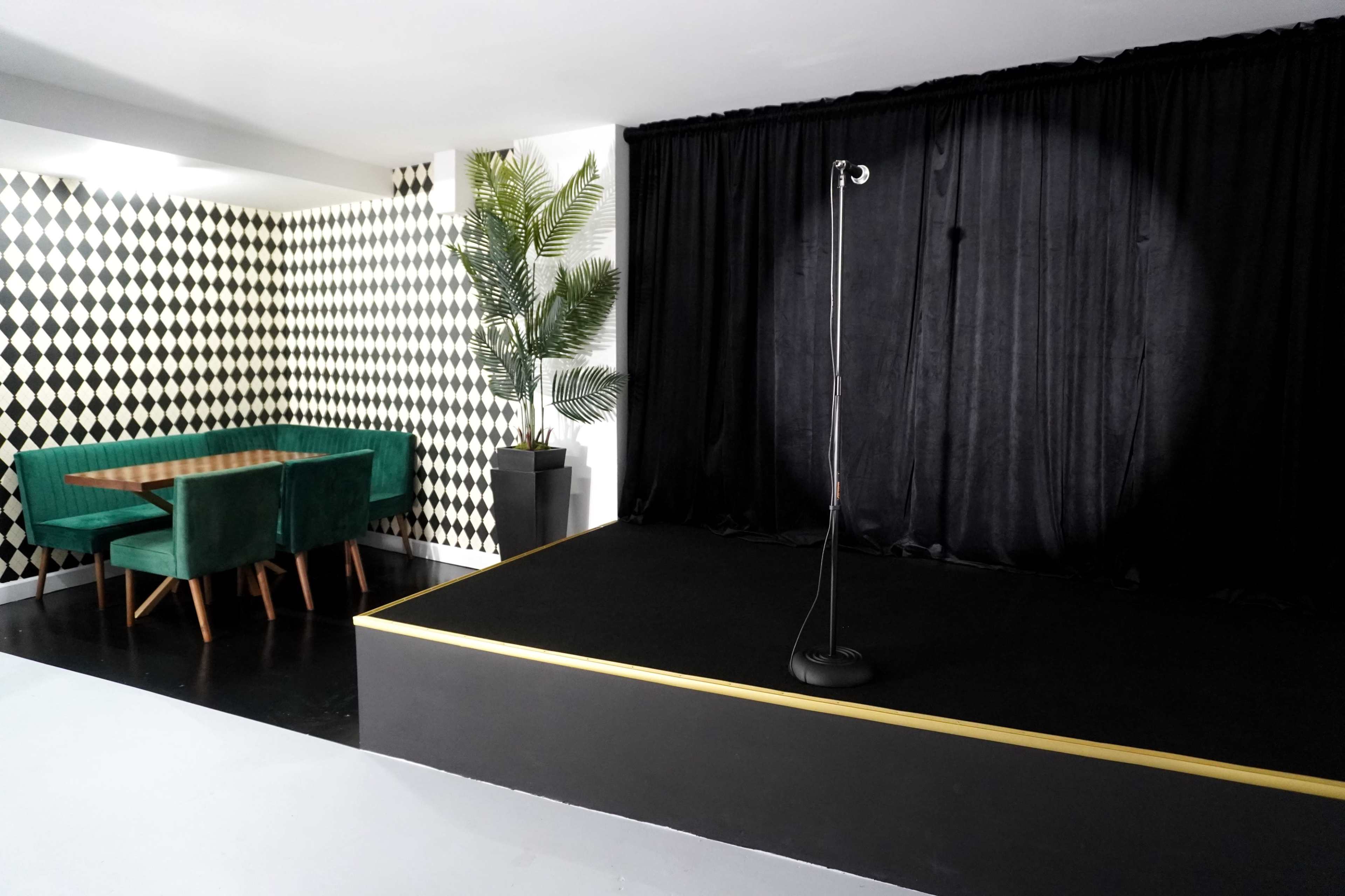 The image shows a small stage with a microphone stand in front of a black curtain, adjacent to a dining area featuring a green velvet table and chairs against a checkered wall.