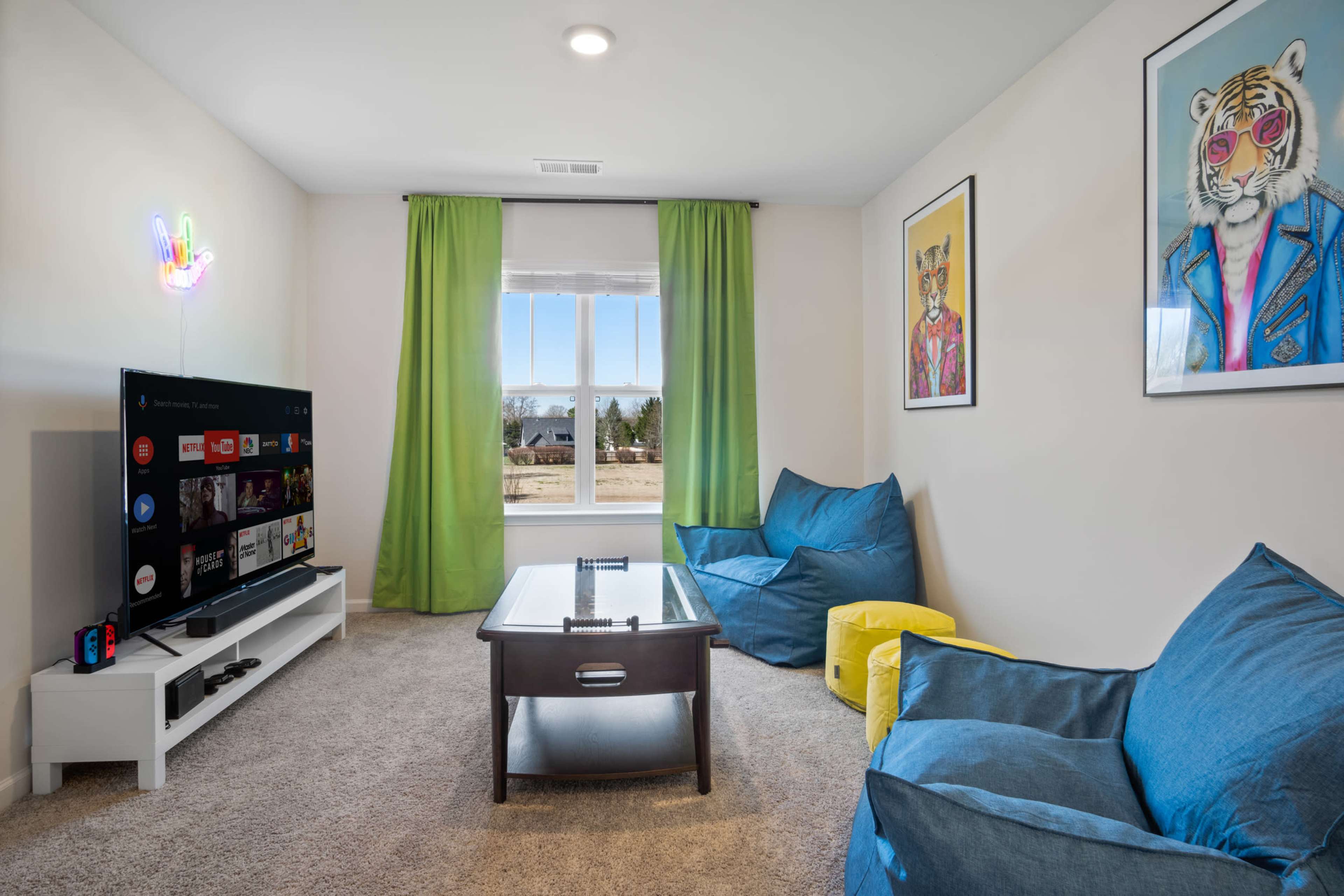 The living room features a large television on a white stand, a dark wooden coffee table, and colorful bean bag chairs near a window with green curtains.