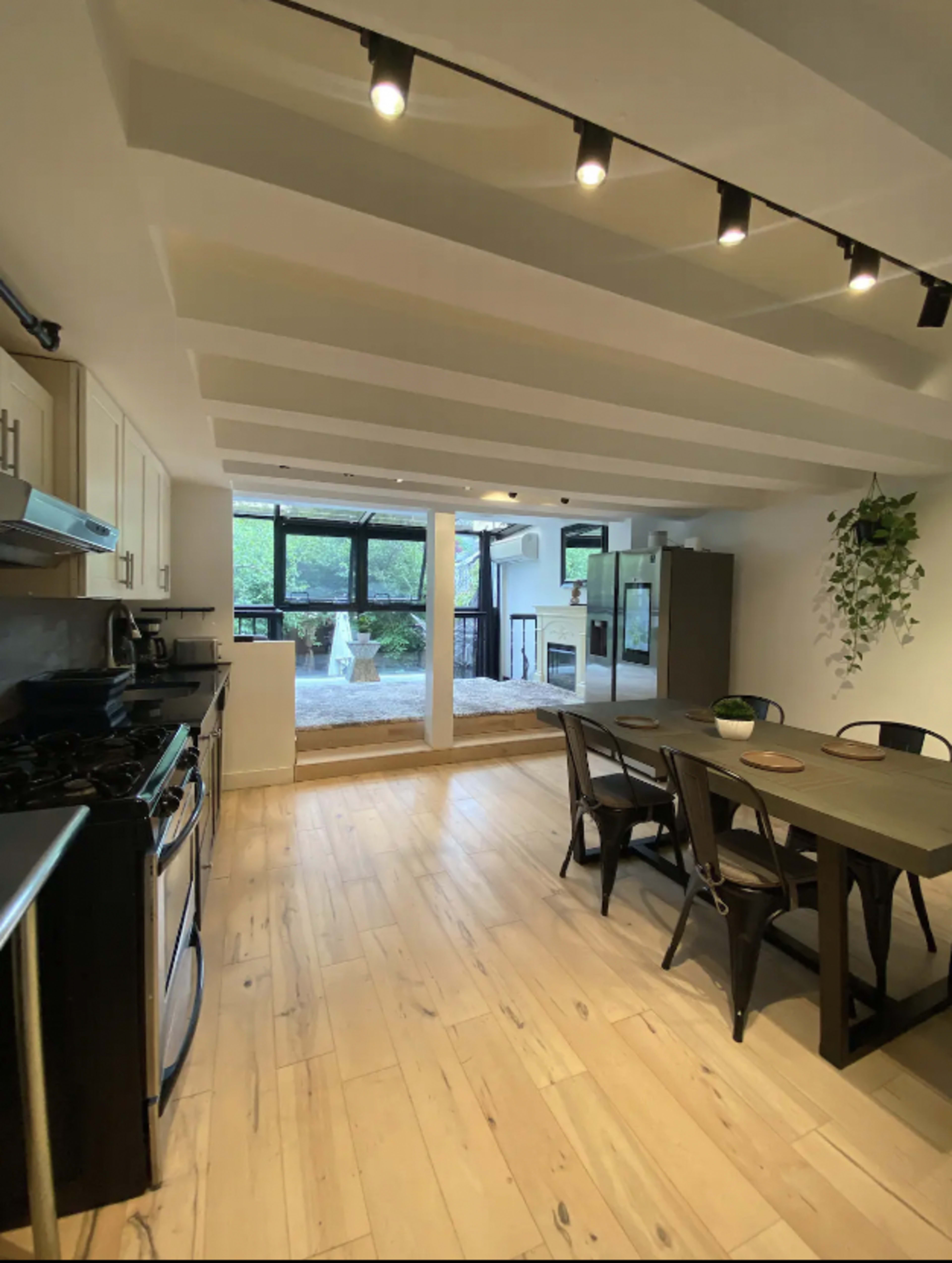 The image shows a modern kitchen and dining area with light wood flooring, featuring a black stove, a dining table with chairs, and large windows overlooking an outdoor space.