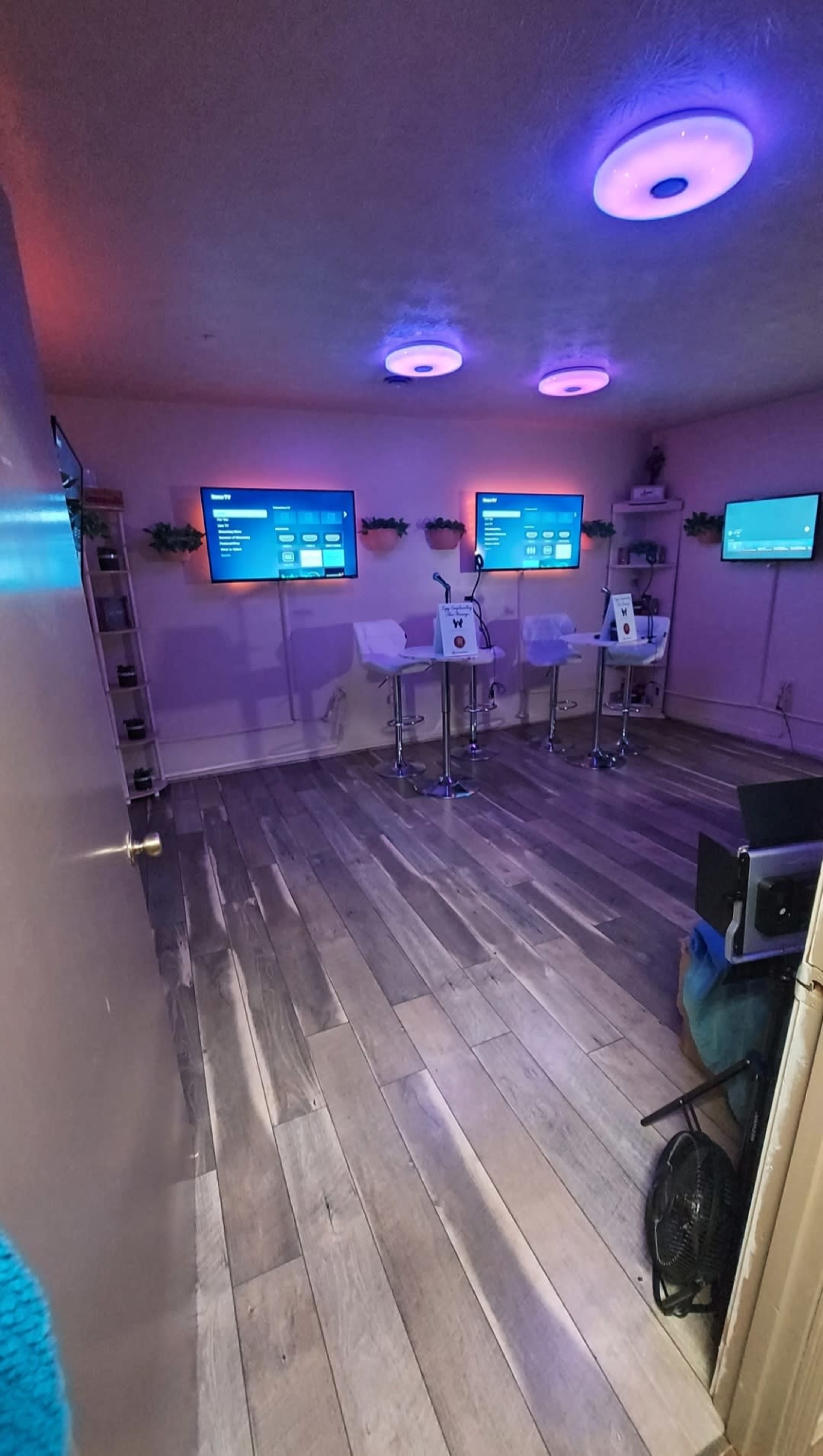 The room features two wall-mounted screens and three high chairs at a table, all set against a floor with wooden planks.