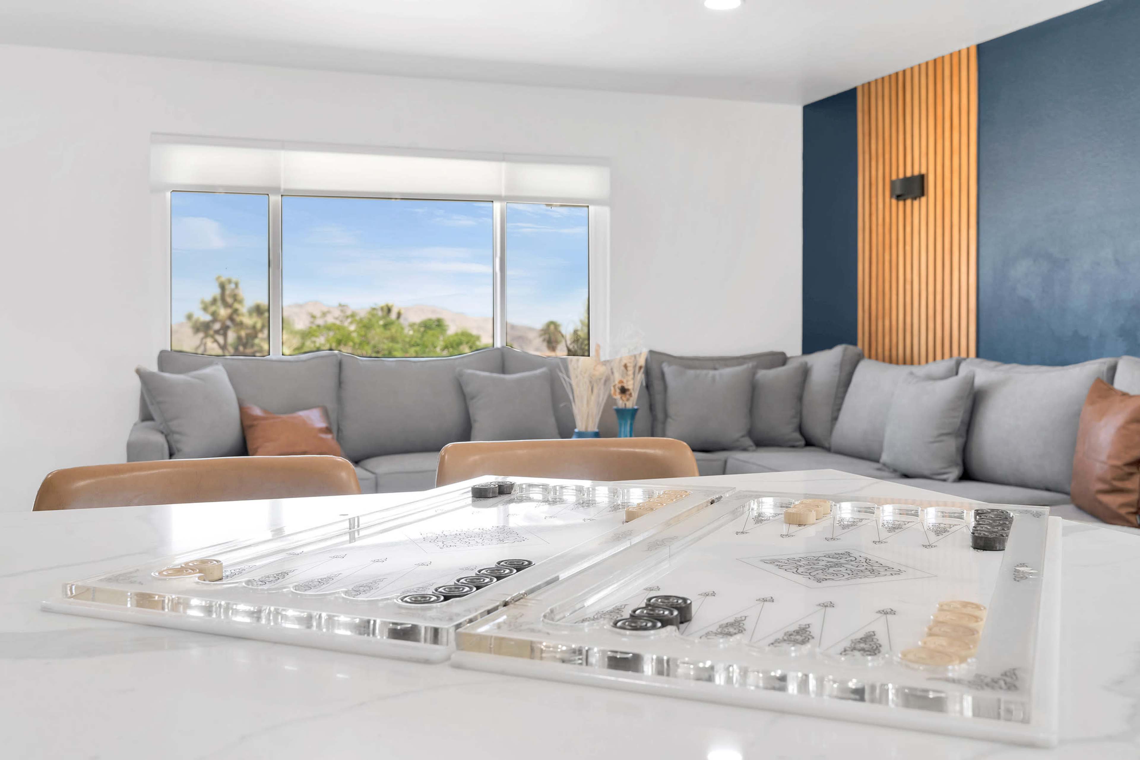 A game board is positioned on a marble table in front of a spacious seating area with gray cushions and a window showcasing an outdoor view.