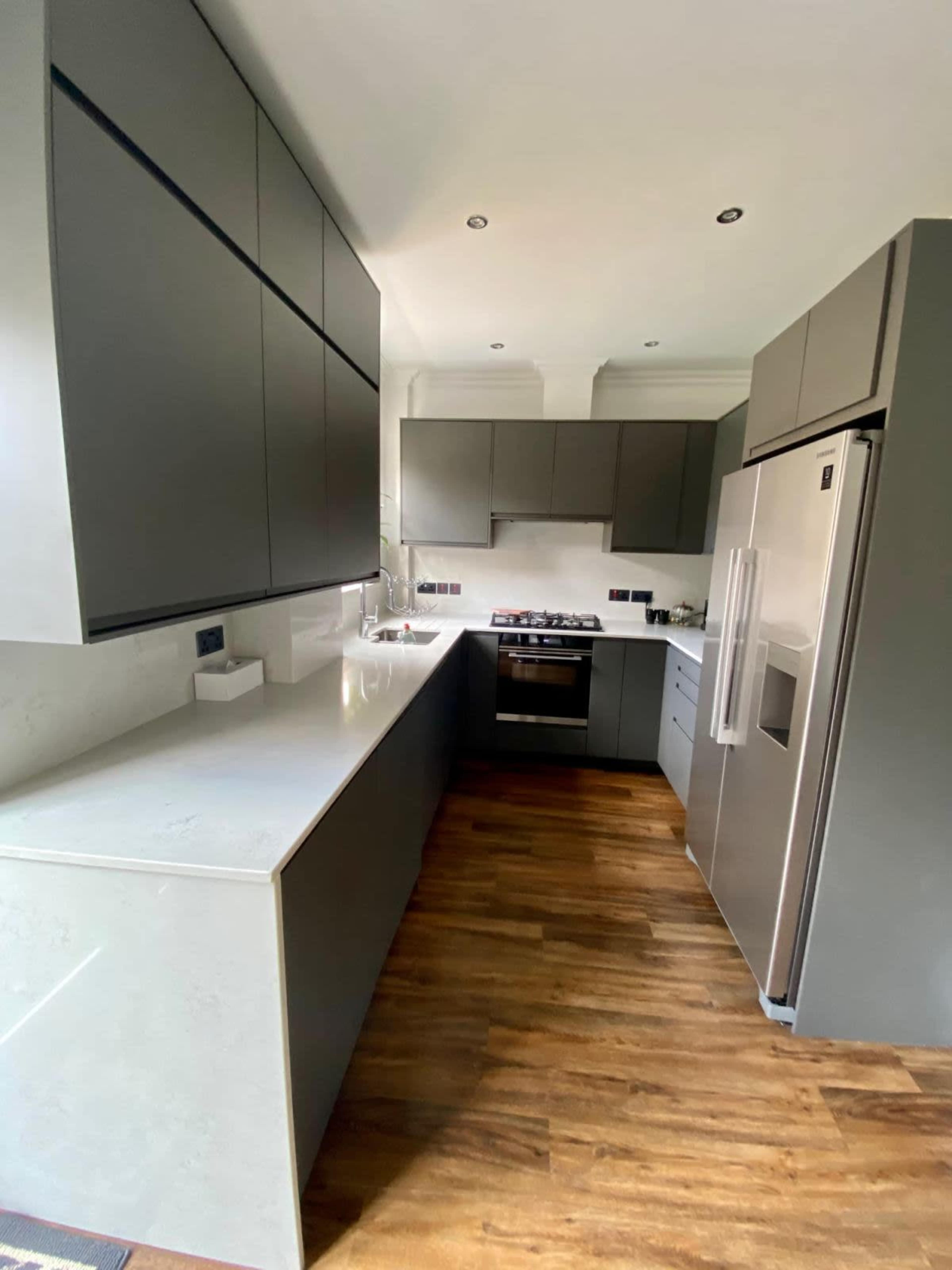 A modern kitchen features gray cabinets, a stainless steel fridge, and a gas stove, with a light-colored countertop and wooden flooring.