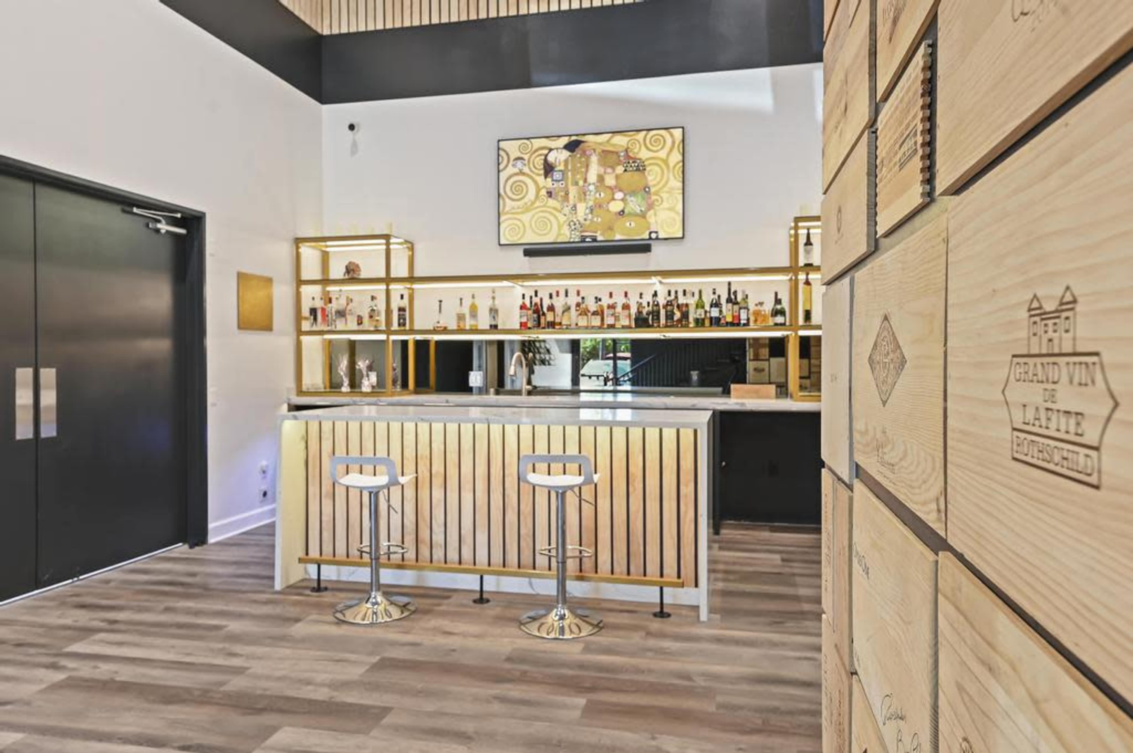 A modern bar area featuring a sleek counter with two stools, a display of various liquor bottles, and a mounted television above.