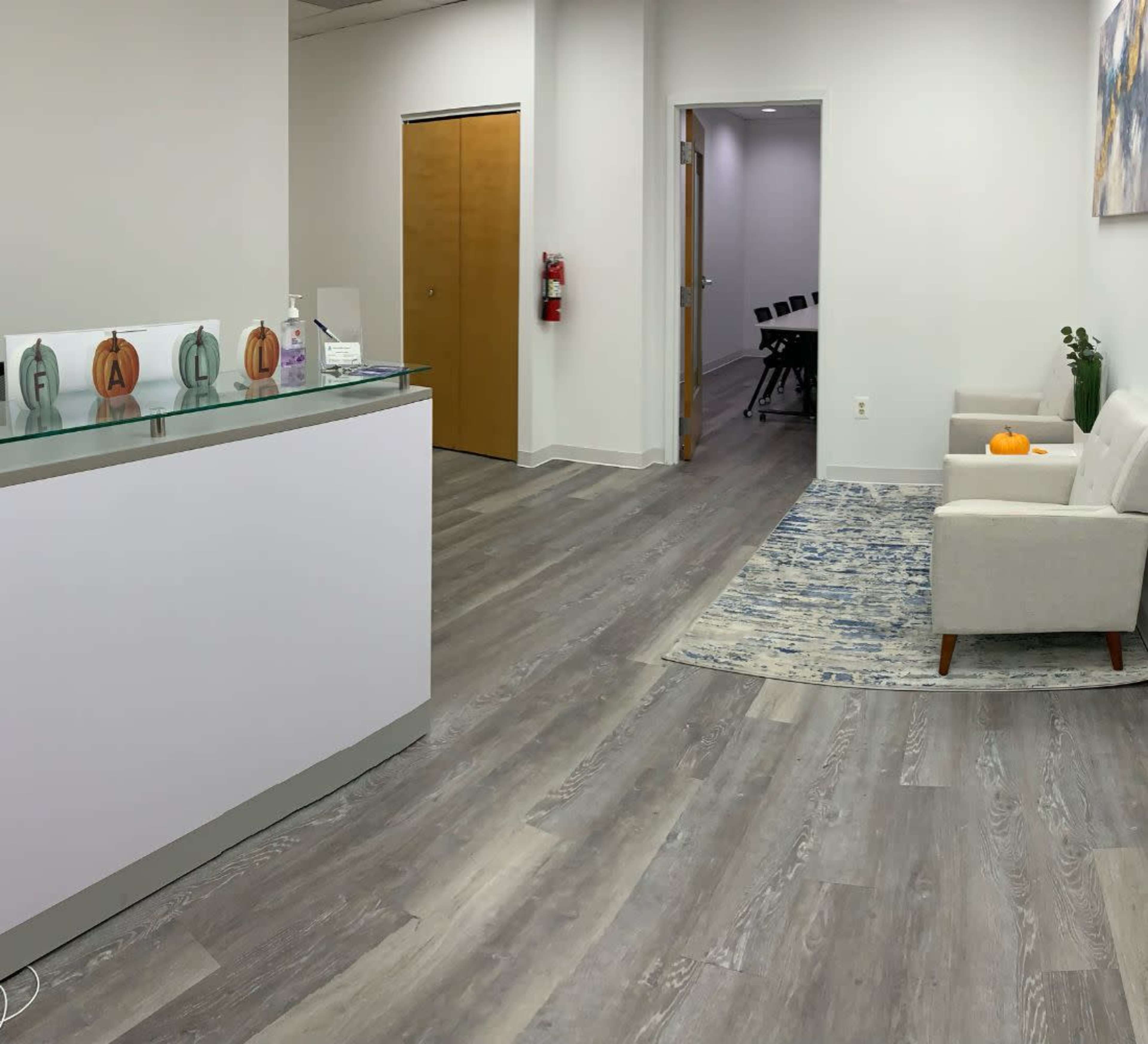A reception area features a white front desk, seating with a small table, and a hallway leading to a meeting room.