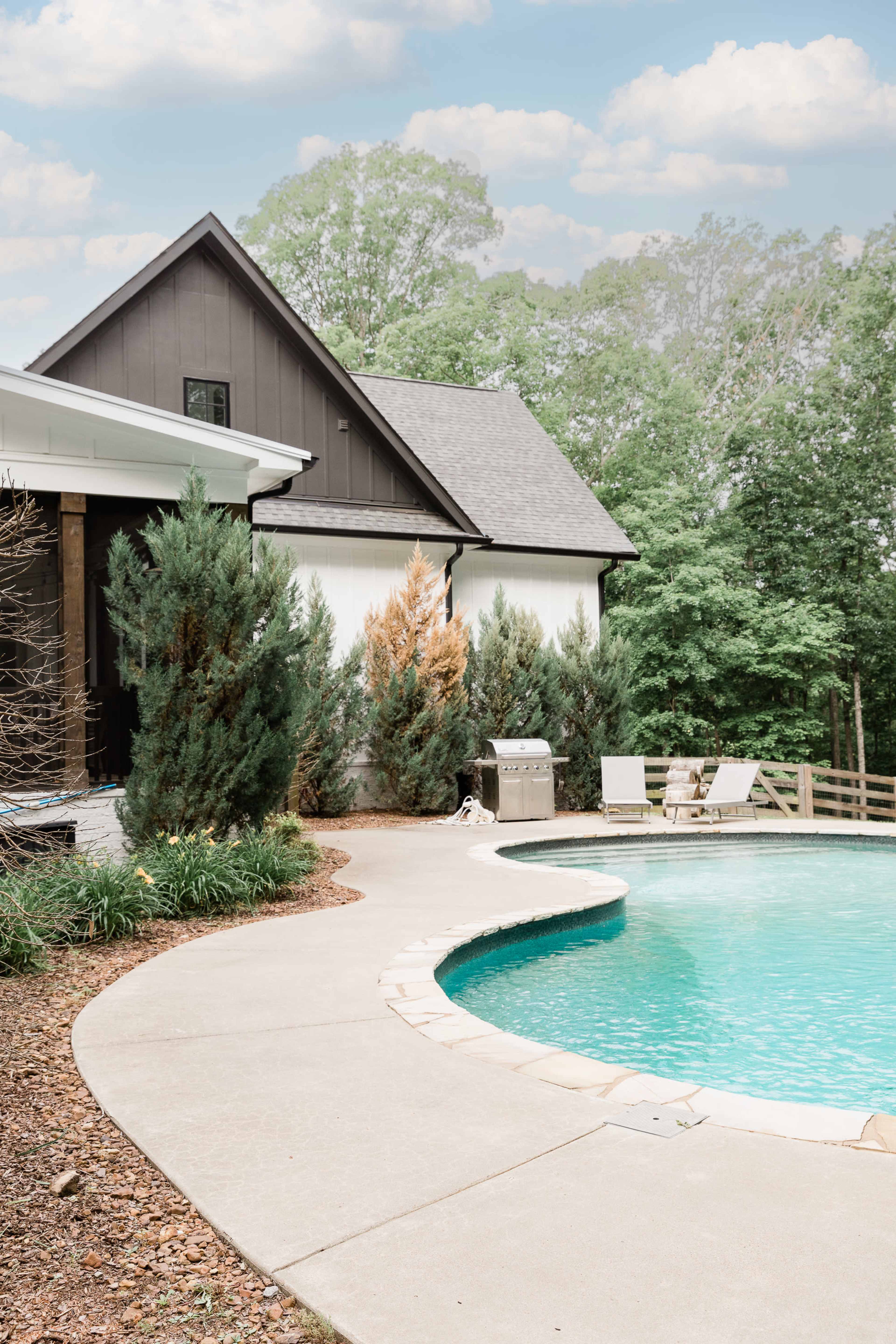 A curved concrete patio surrounds a swimming pool next to a modern house with a dark exterior and lush greenery.