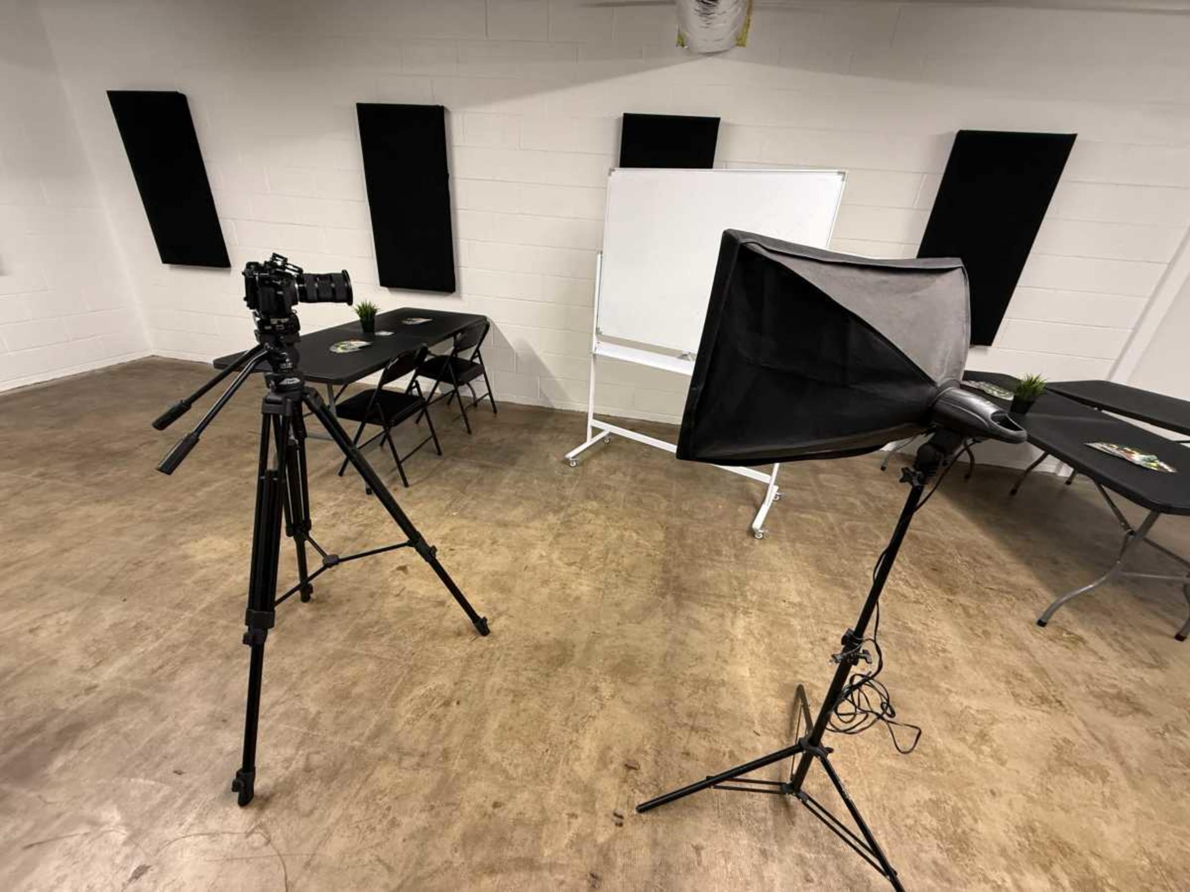 A camera on a tripod and a lighting setup are positioned in a room with black acoustic panels and a whiteboard.