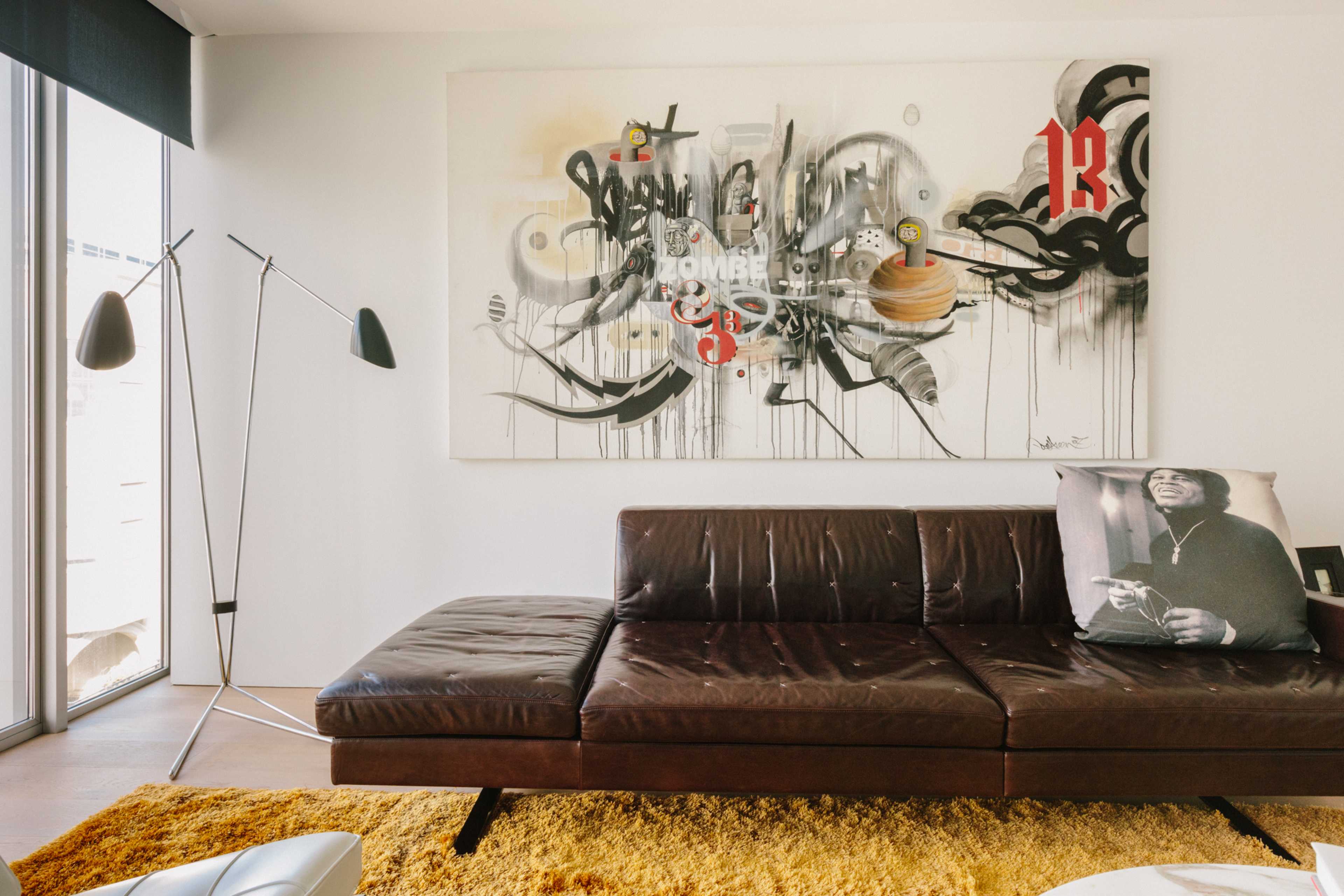 A modern living room features a dark leather sectional sofa, a large abstract painting on the wall, and a standing lamp beside a vibrant yellow rug.