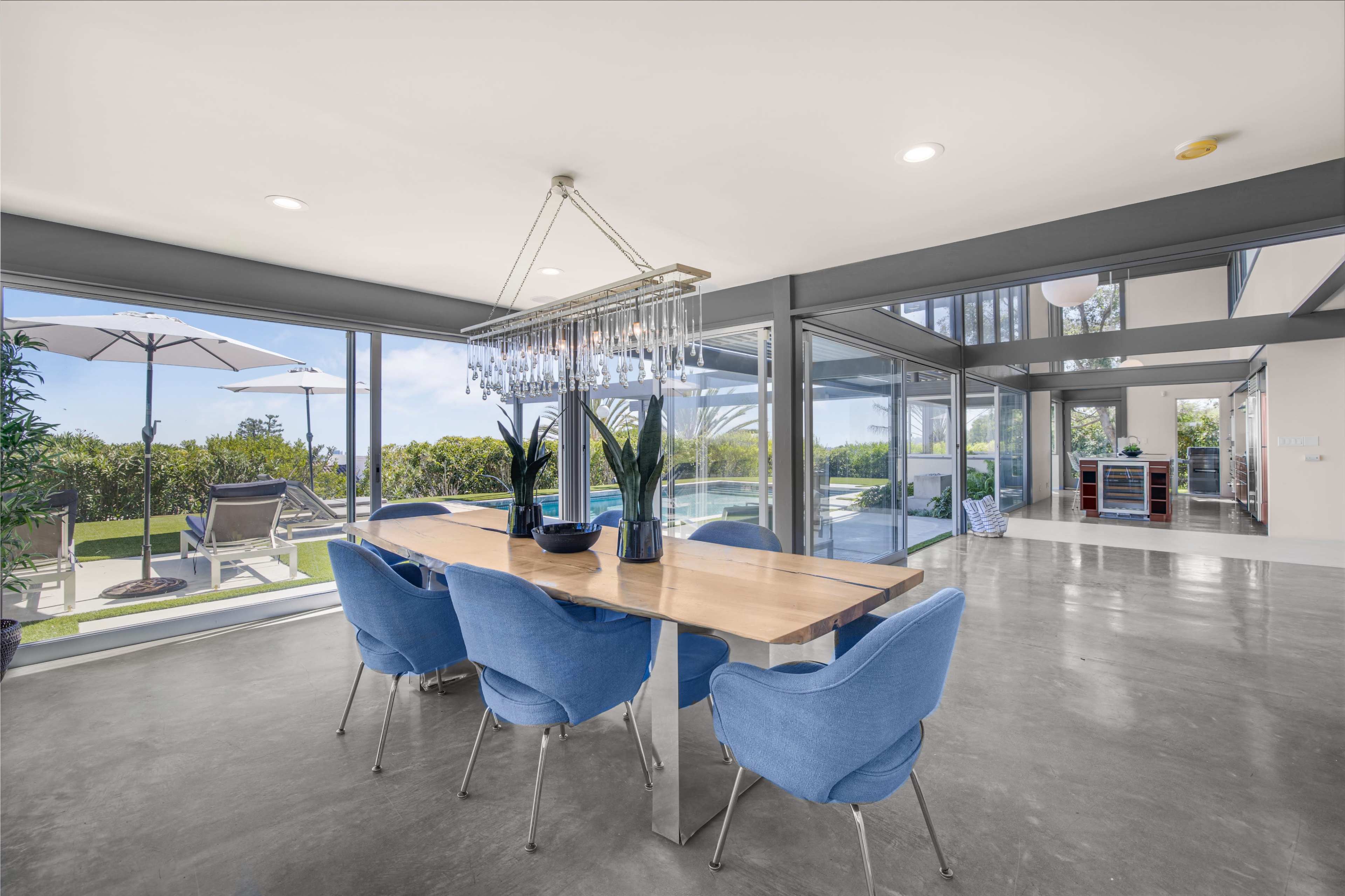 A modern dining area features a long wooden table surrounded by blue chairs, with large glass windows offering a view of an outdoor space.