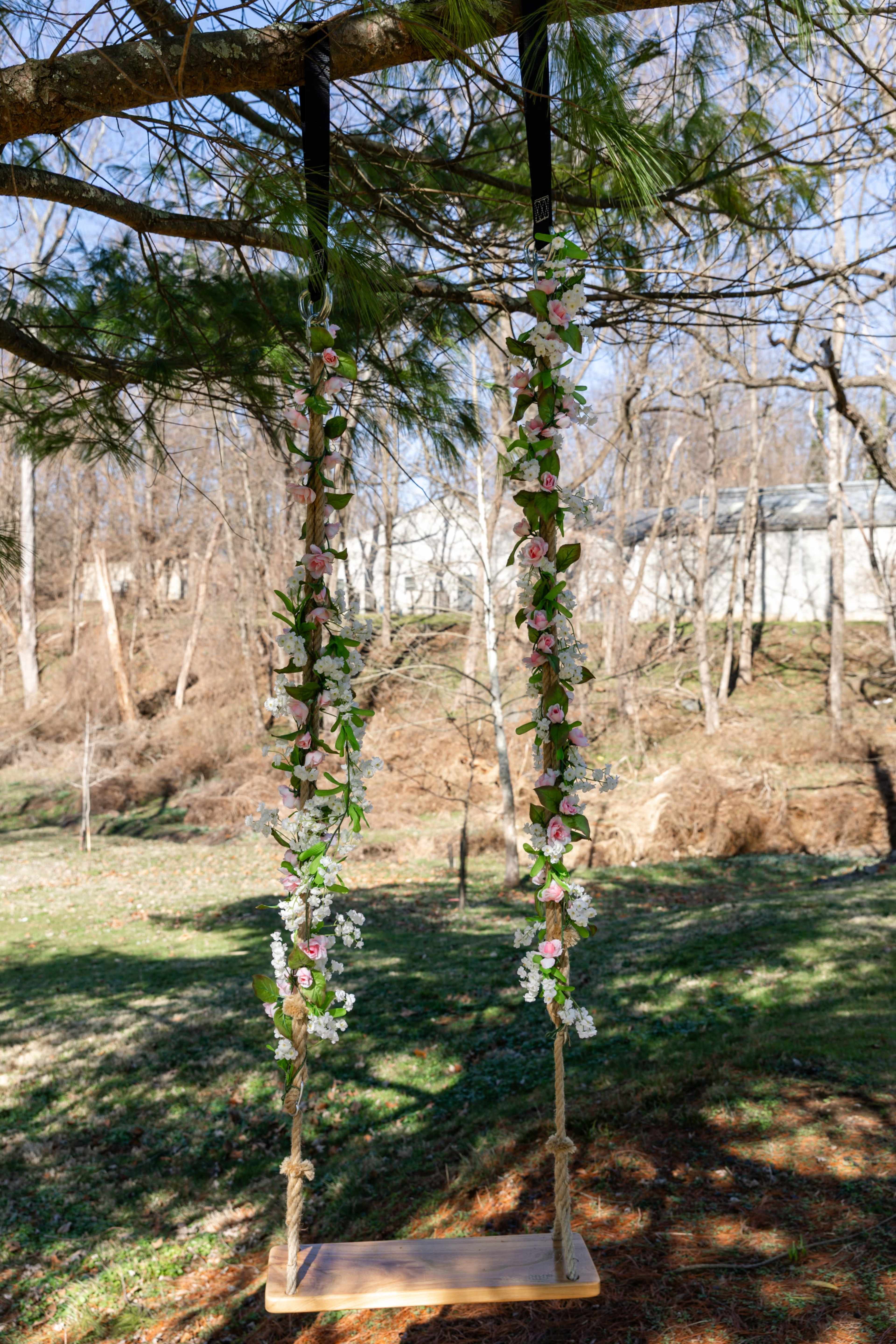 A wooden swing adorned with flowers hangs from a tree branch in a grassy yard.