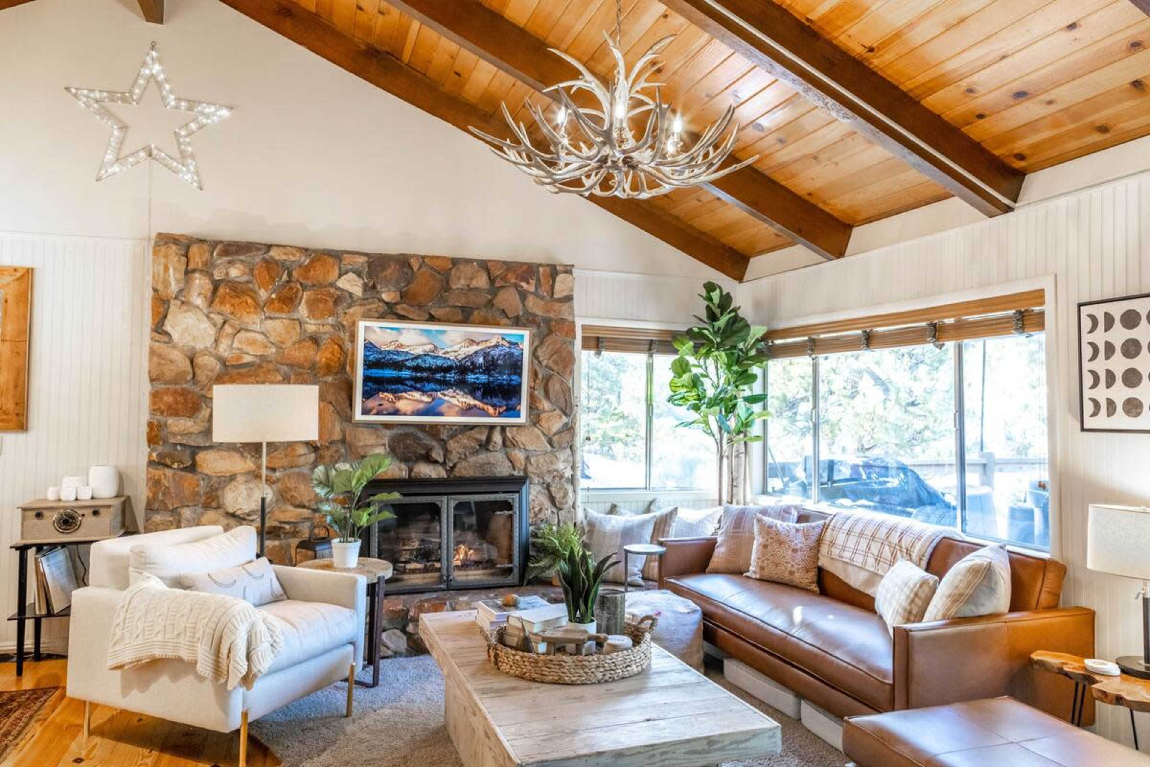 The image shows a cozy living room with a stone wall fireplace, wooden beams, a large window, and comfortable seating arrangements.