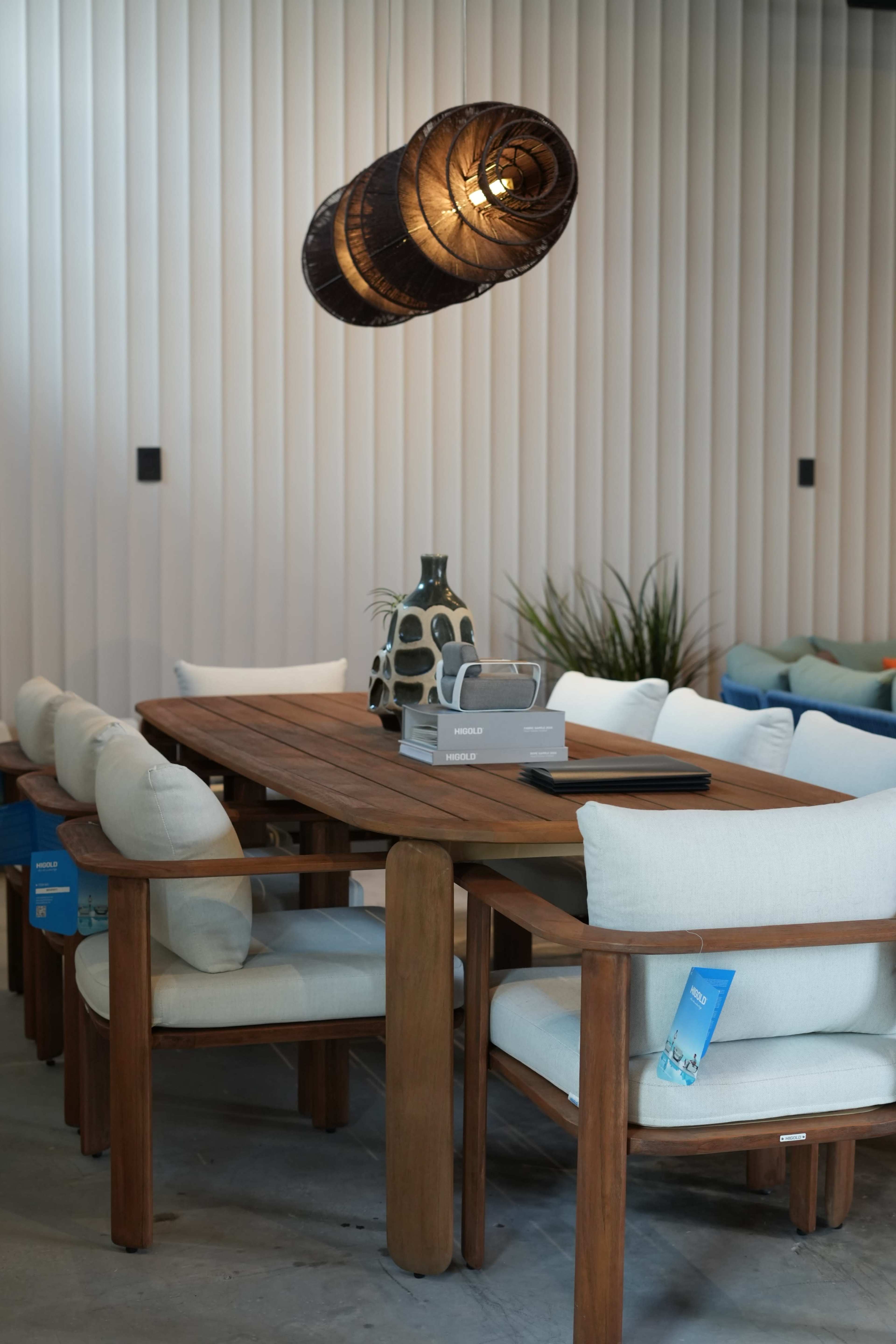 A wooden dining table surrounded by light-colored upholstered chairs is illuminated by a pendant light, with a decorative vase on the table and various plants in the background.
