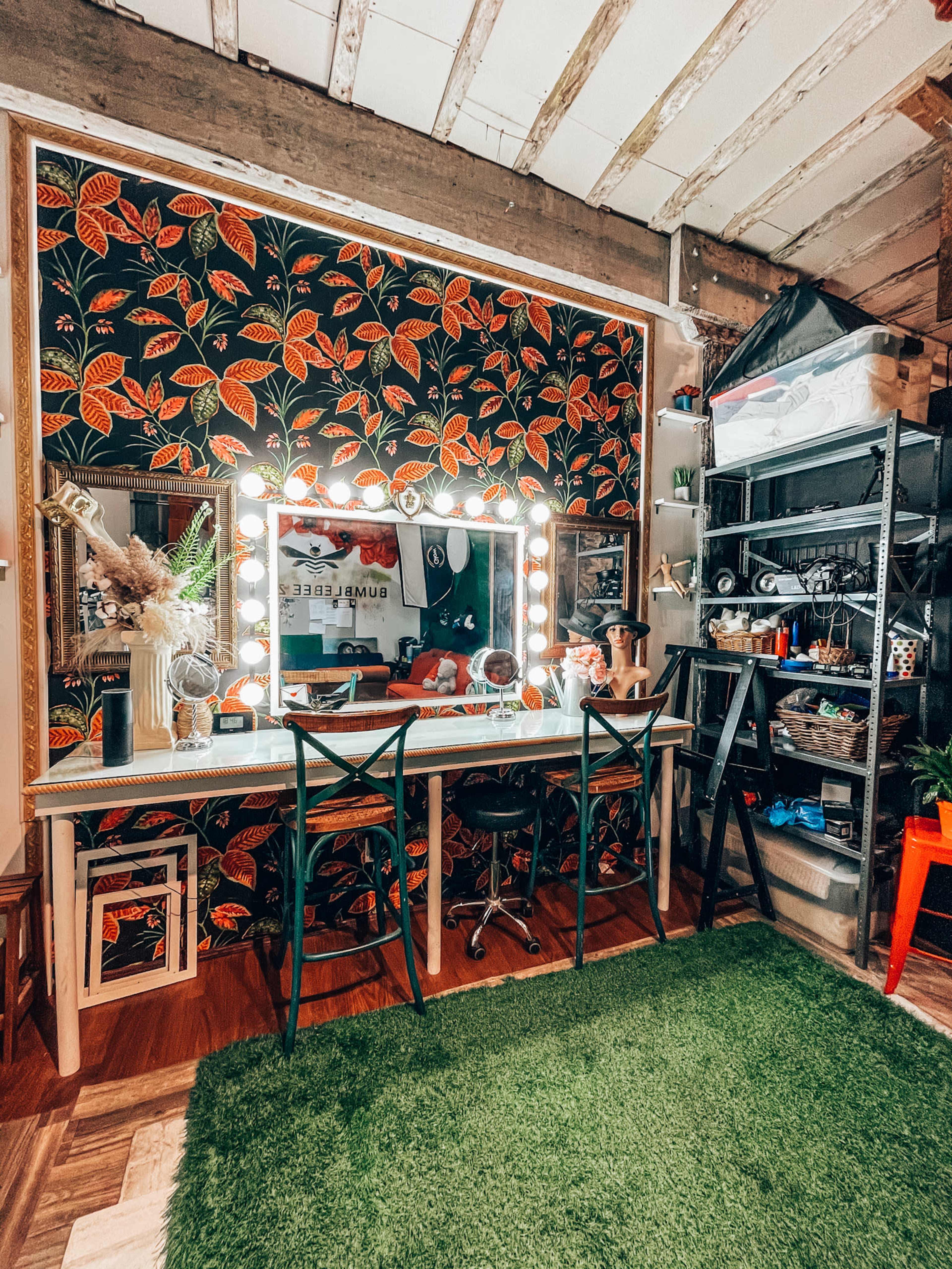 A brightly lit vanity area features a large mirror surrounded by lights, set against a floral-patterned wall, with a green rug on the floor and various storage units nearby.