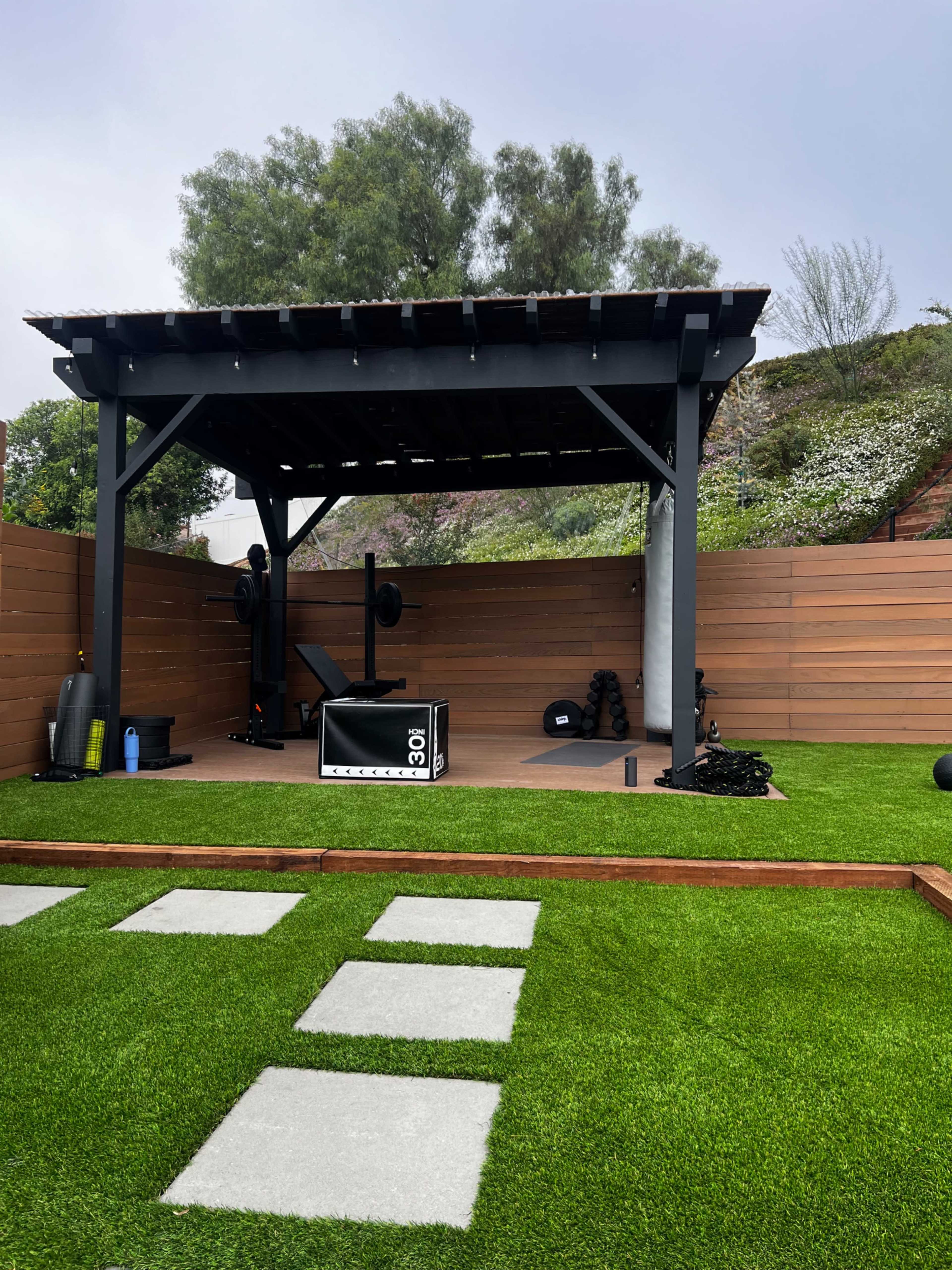 The image shows a backyard gym area with a covered workout space featuring exercise equipment, surrounded by a well-maintained lawn and wooden fencing.