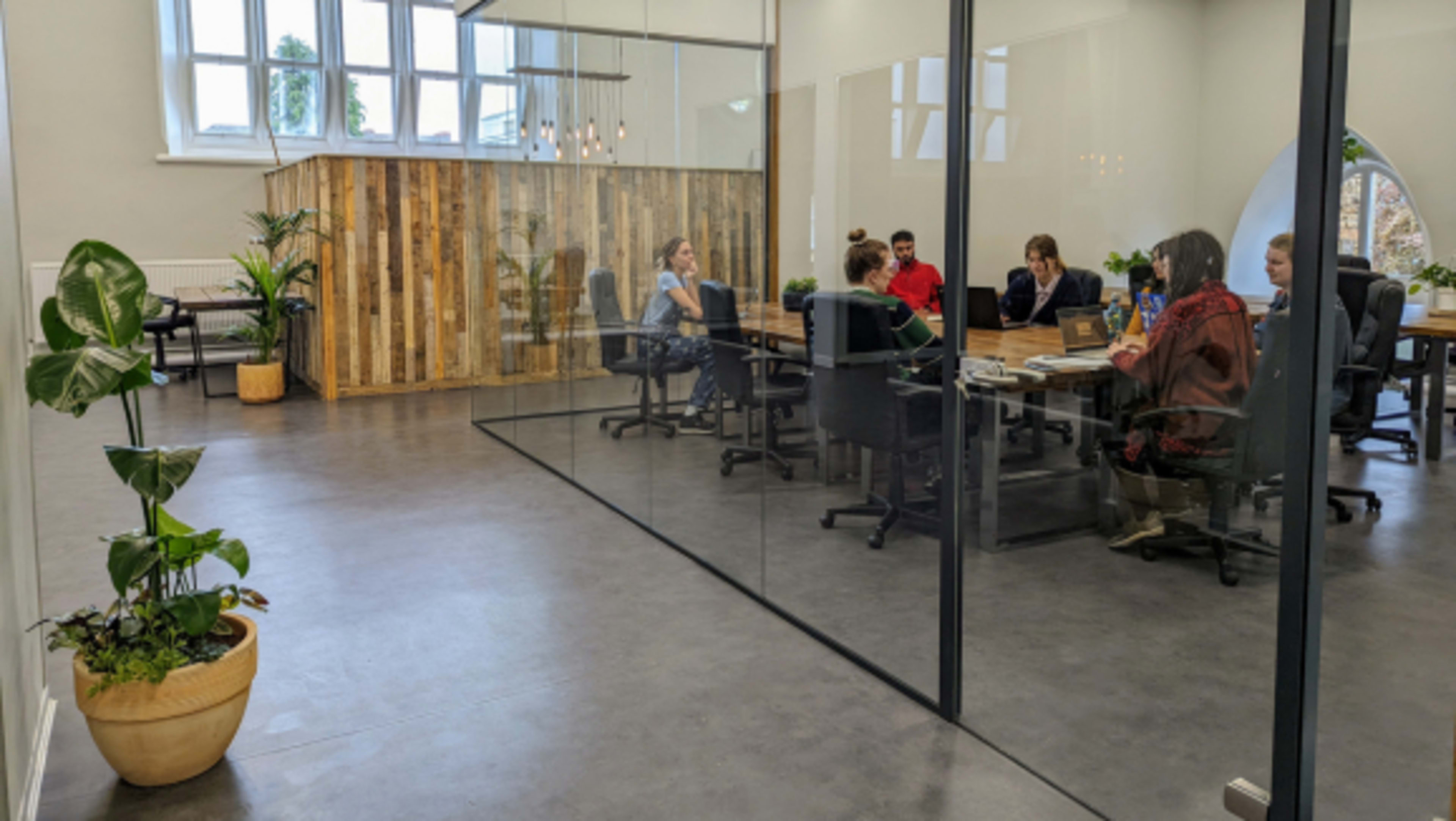 A group of people sit around a large table in a modern office space with glass walls and wooden accents.
