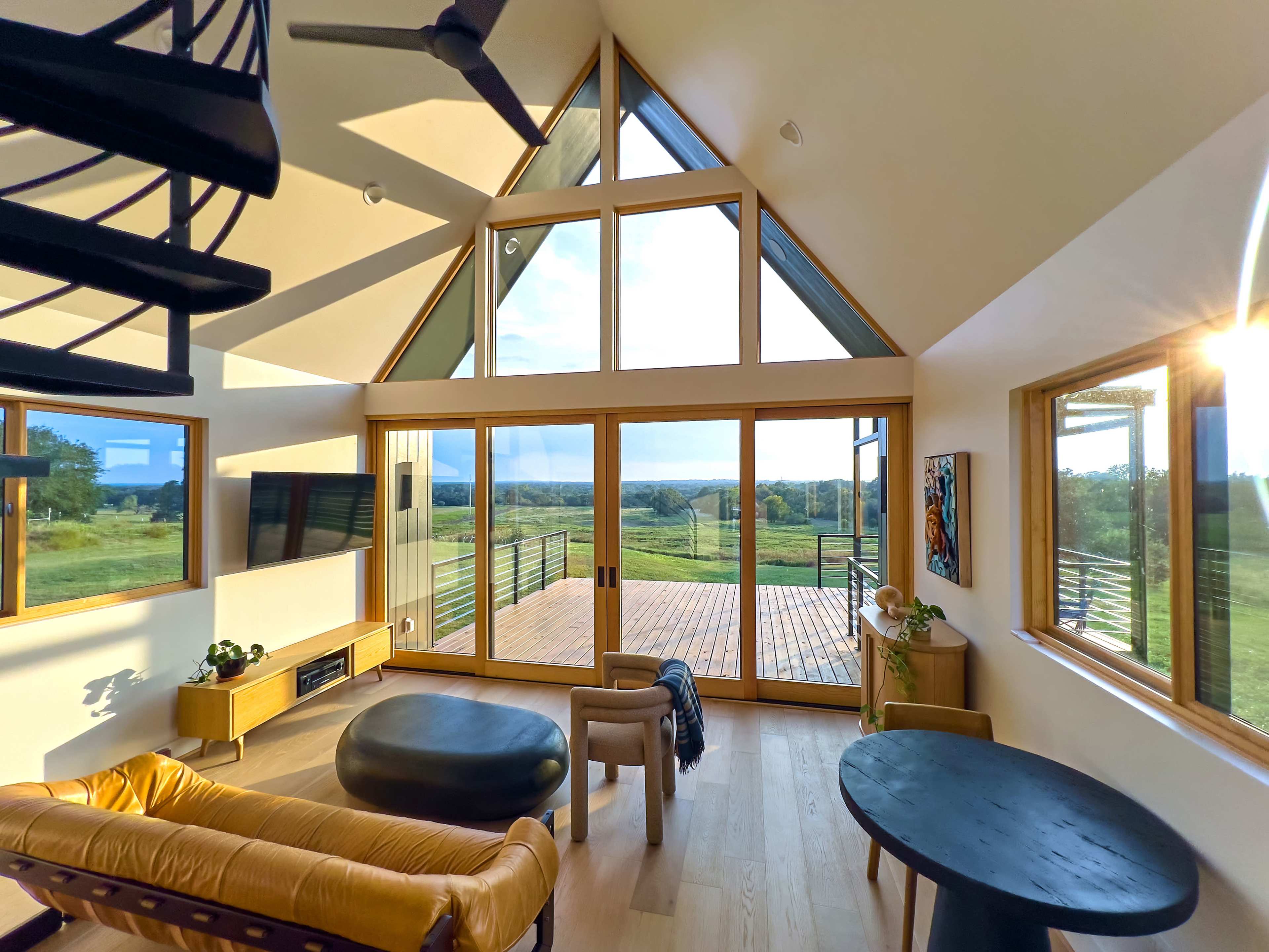 The image shows a modern living space with large triangular windows, wooden flooring, and furniture arranged to face a deck overlooking a green landscape.