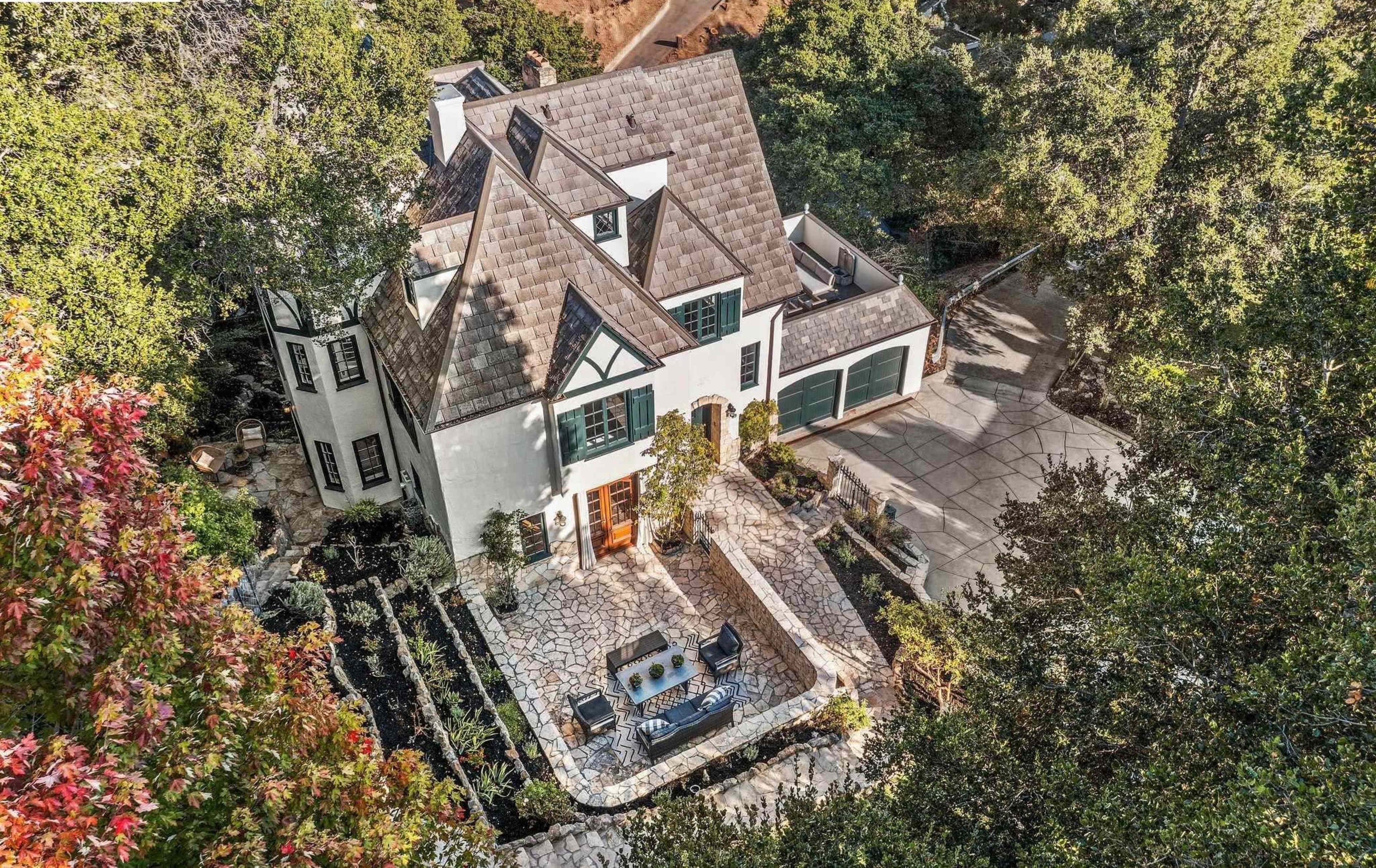 The image shows a large, two-story house with a steeply pitched roof, surrounded by trees and featuring a stone patio in front.