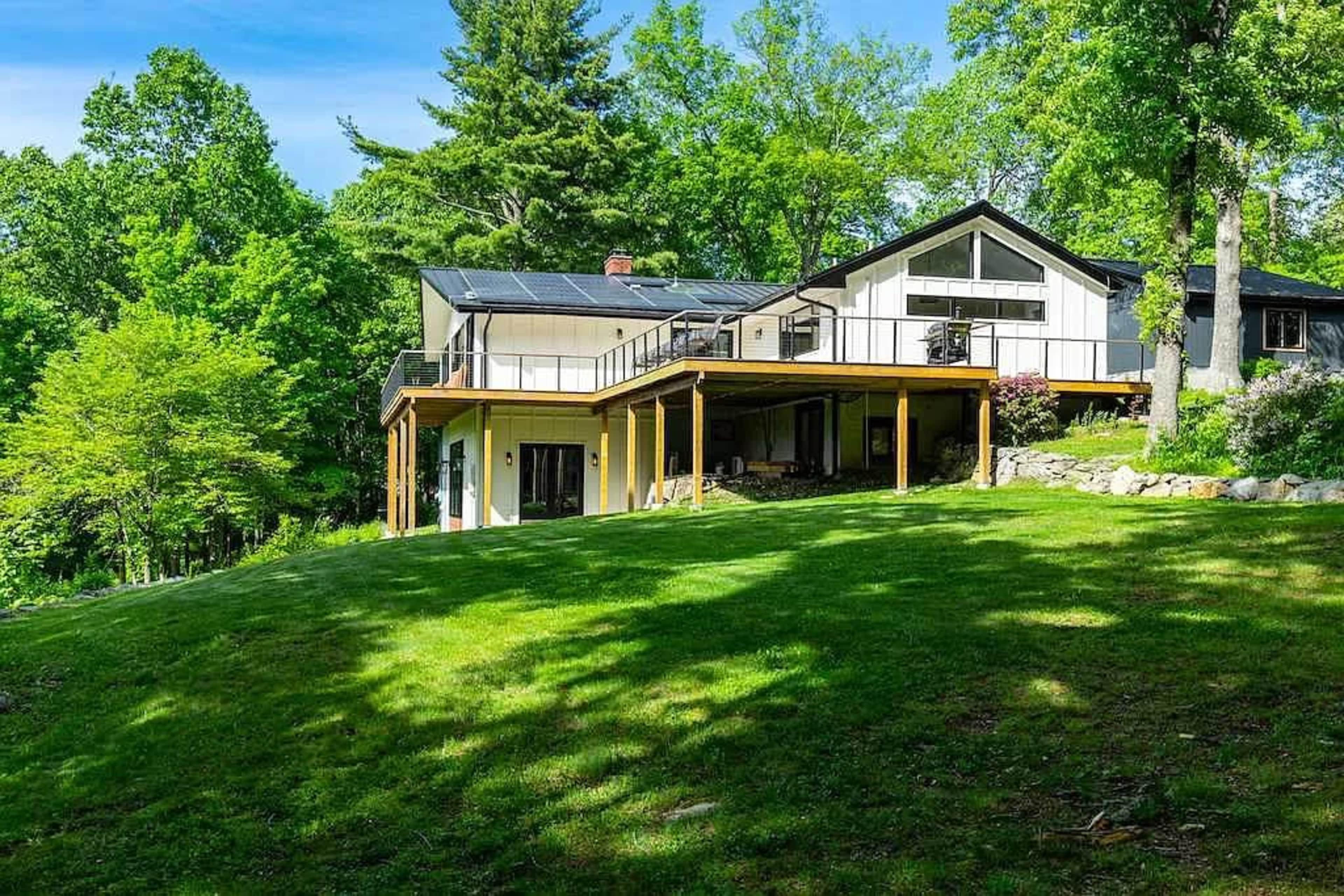 A modern house with large windows and a deck overlooks a green lawn surrounded by trees.