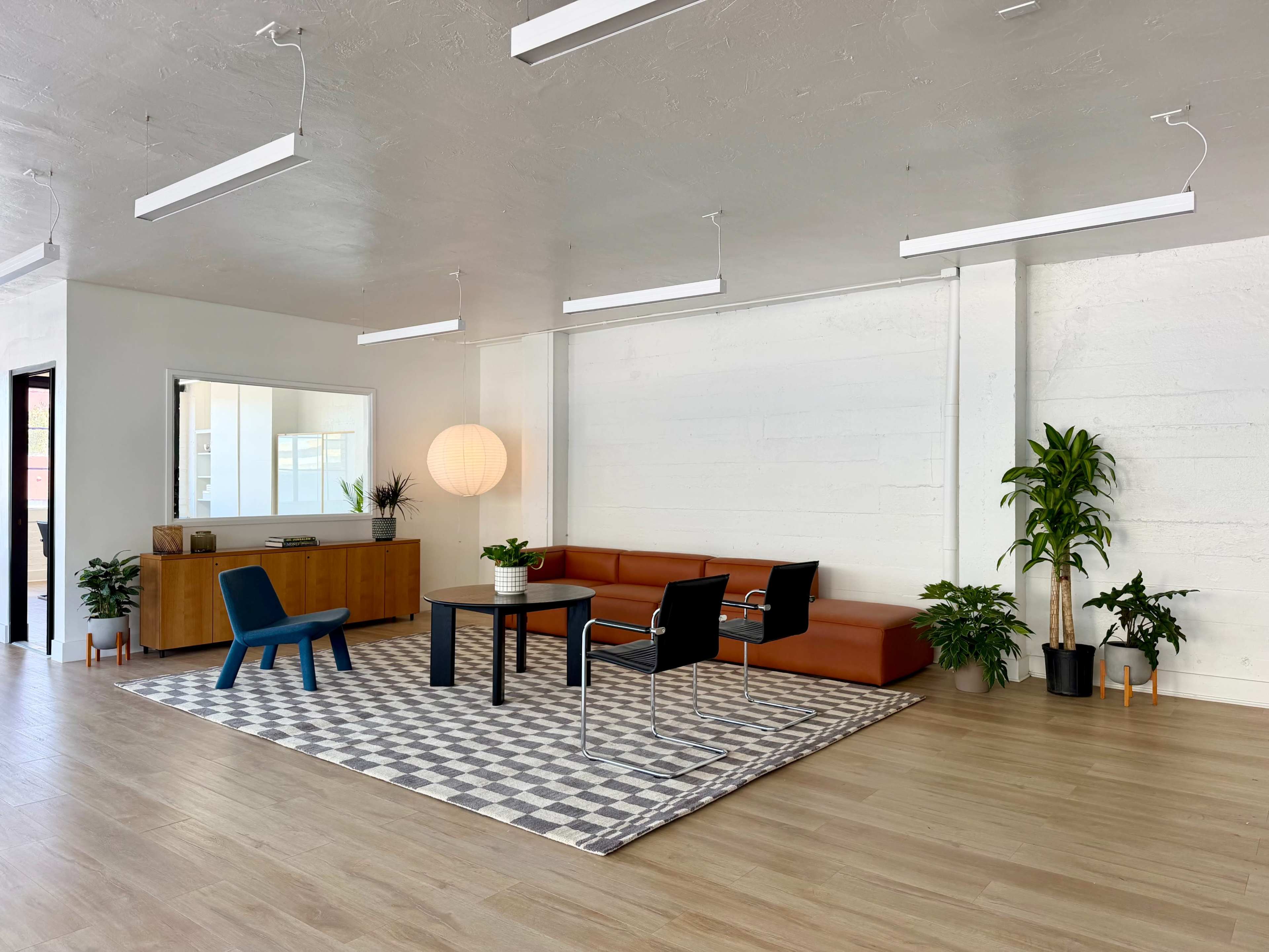 A modern office space features a leather sofa, a wooden cabinet, a round lamp, plants, and a geometric rug with a table and chairs.