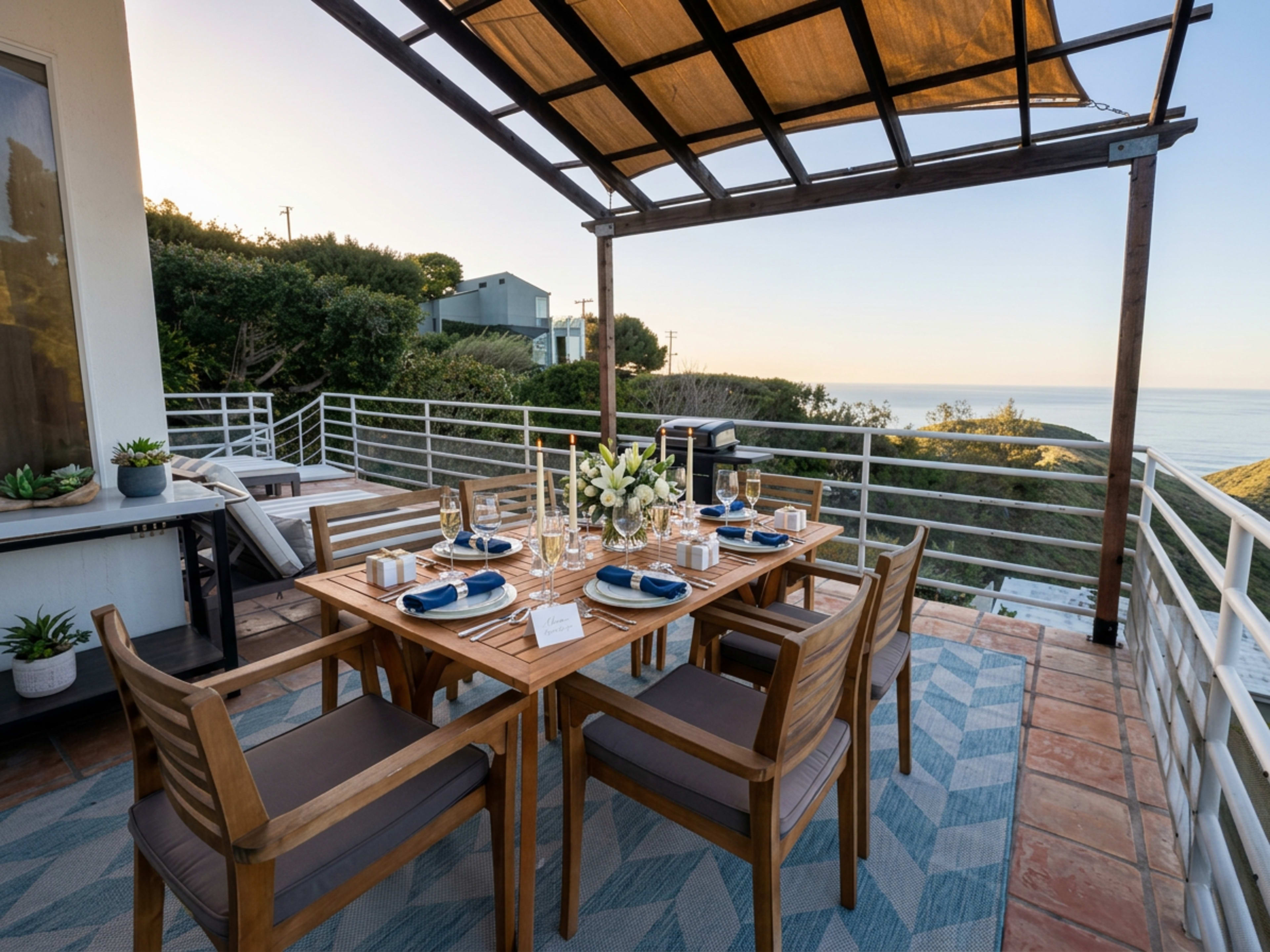 A wooden dining table is set on a patio overlooking the ocean, adorned with plates, glasses, and floral arrangements.