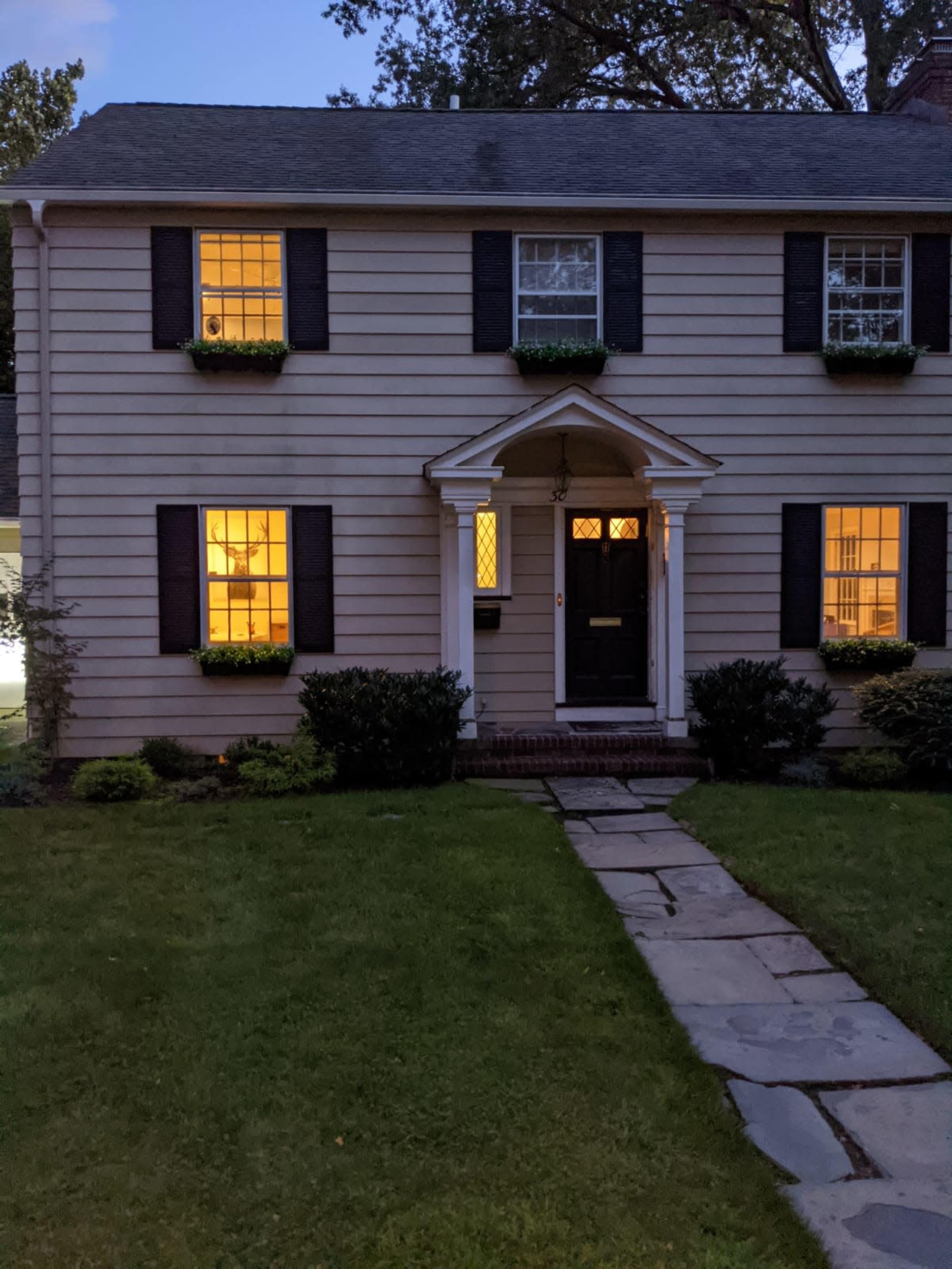 A two-story house with black shutters and illuminated windows has a stone pathway leading to the front door.