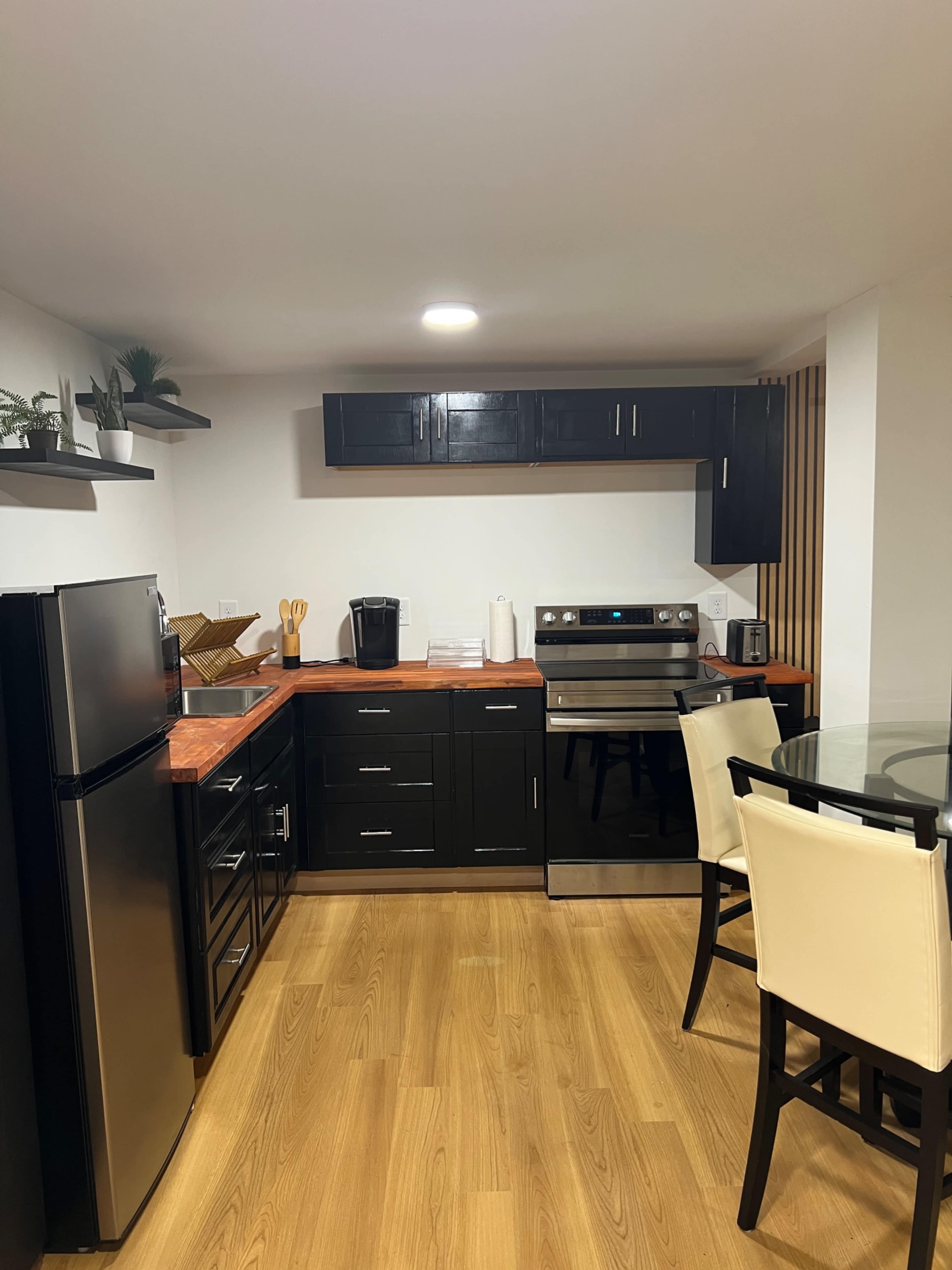 The image shows a compact kitchen with dark cabinets, stainless steel appliances, and a glass dining table.