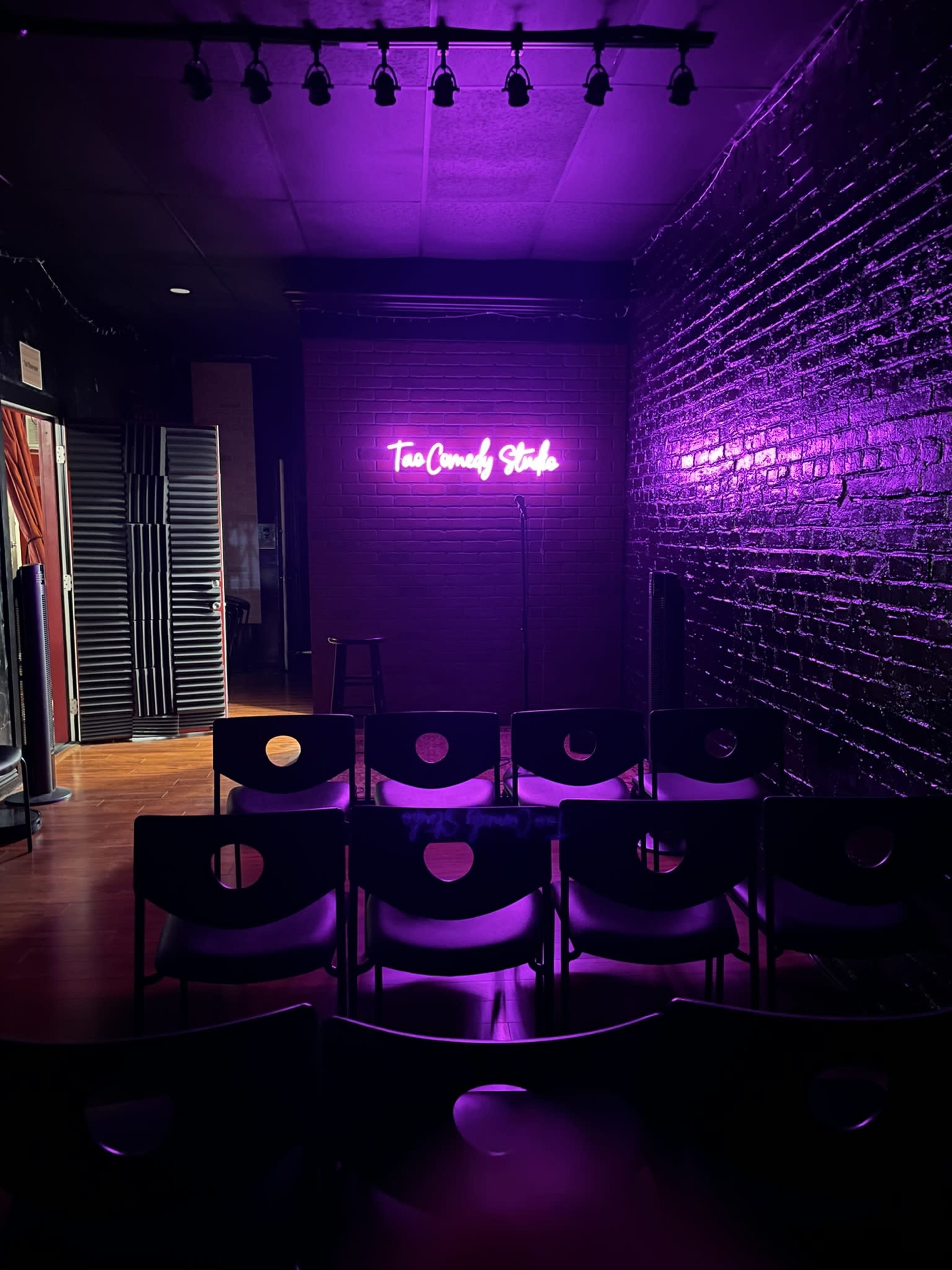 The image shows a dimly lit comedy venue with black brick walls, purple neon signage that reads "The Comedy Stage," and rows of chairs set up for an audience.