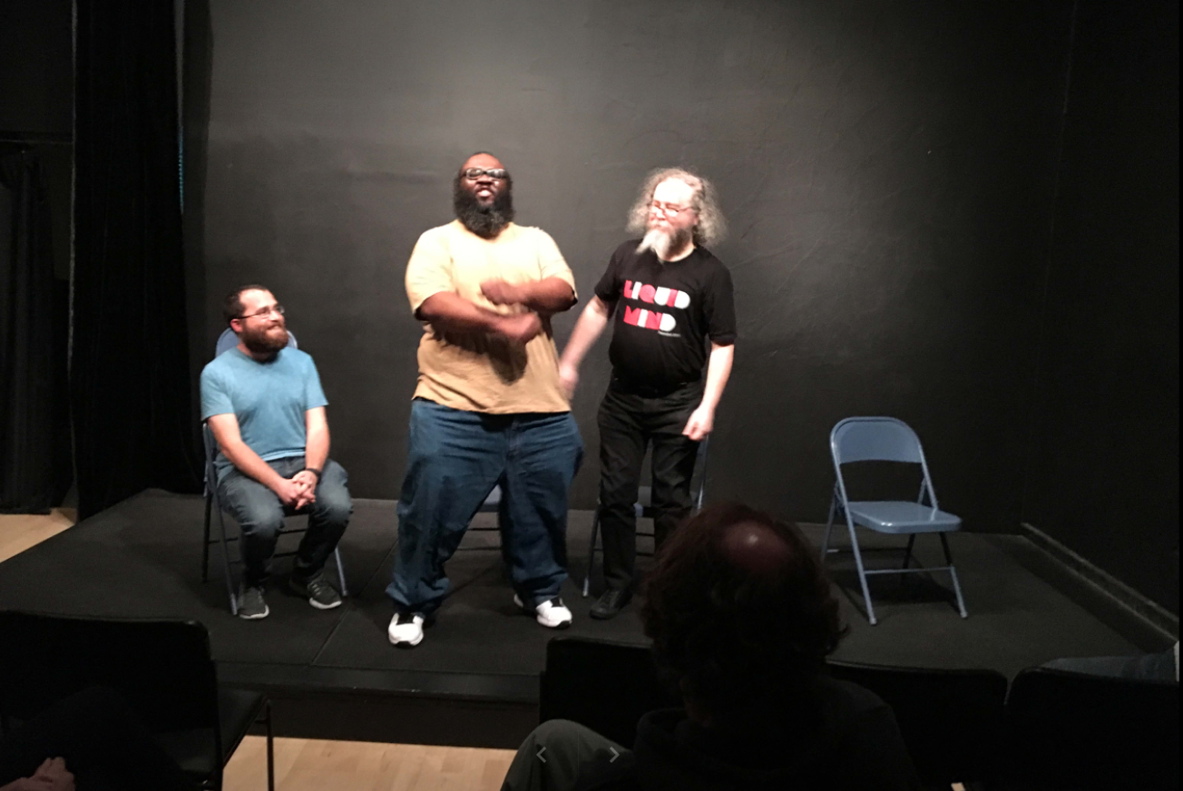 Three men are performing on a small stage, with two standing and one sitting on a chair.