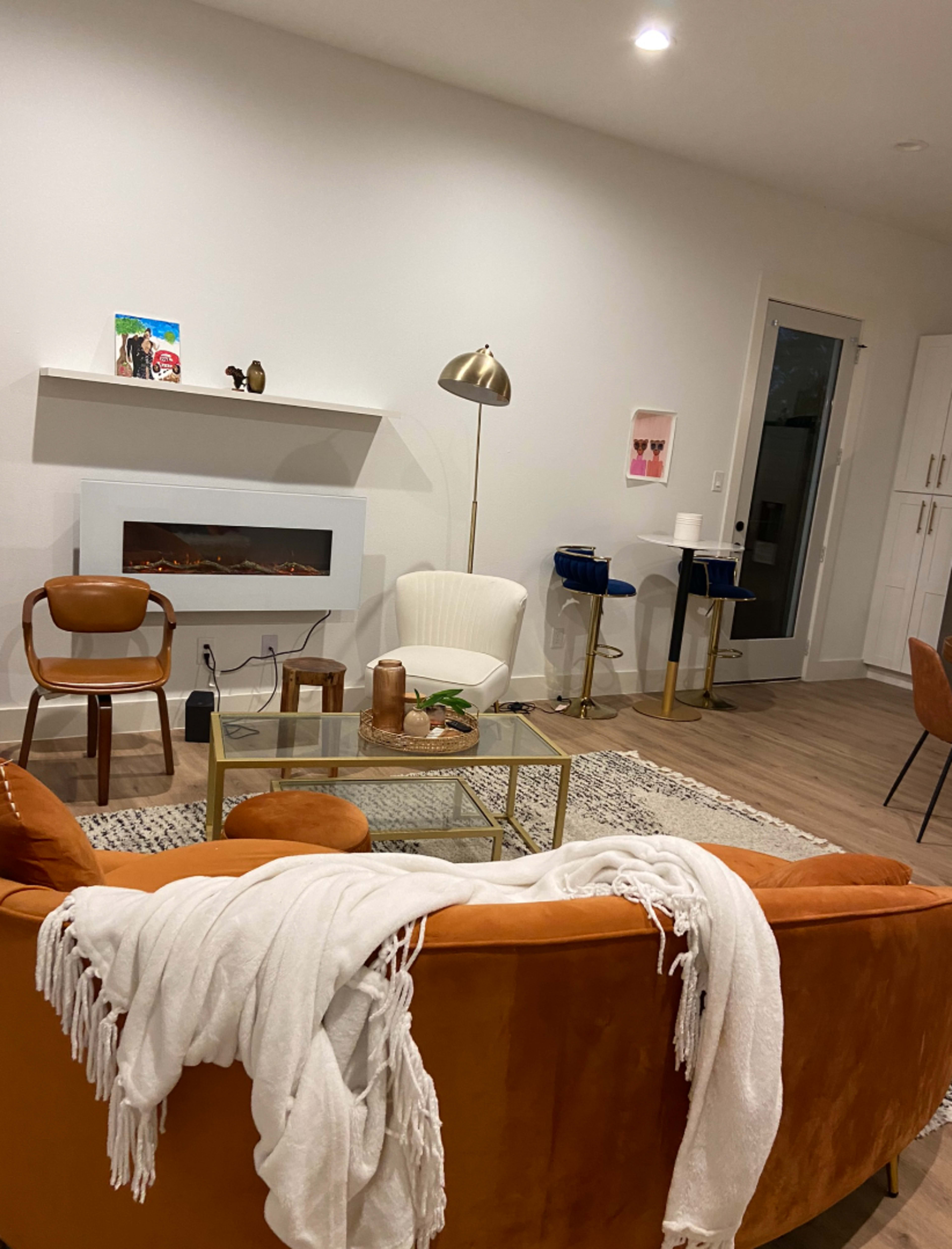 A modern living room with a brown couch, a glass coffee table, and various seating arrangements, including a white armchair and wooden chairs, along with a wall-mounted fireplace.