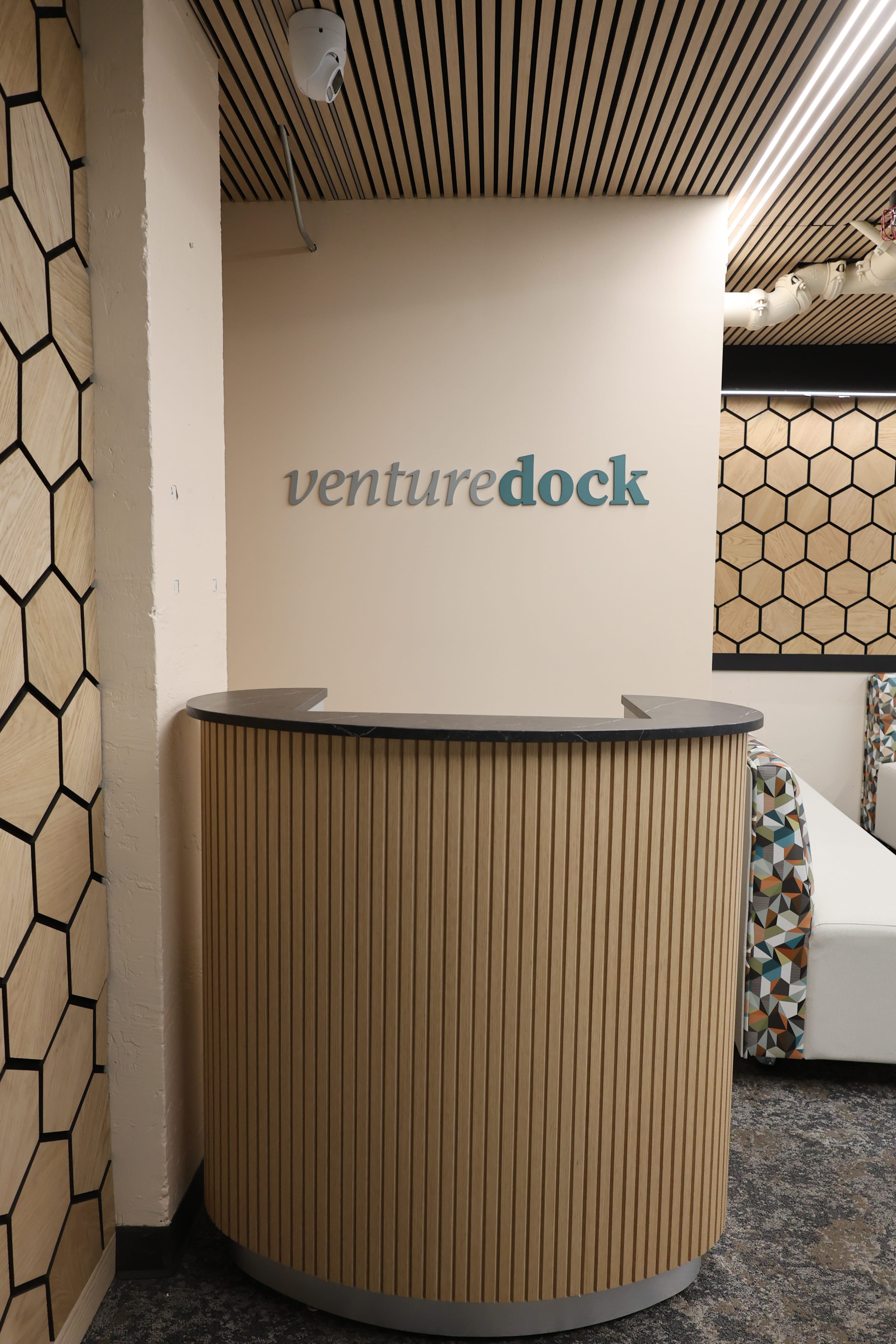 The image shows a modern reception area with a curved wooden desk, featuring the name "venturedock" on the wall behind it, and geometric-patterned decor nearby.