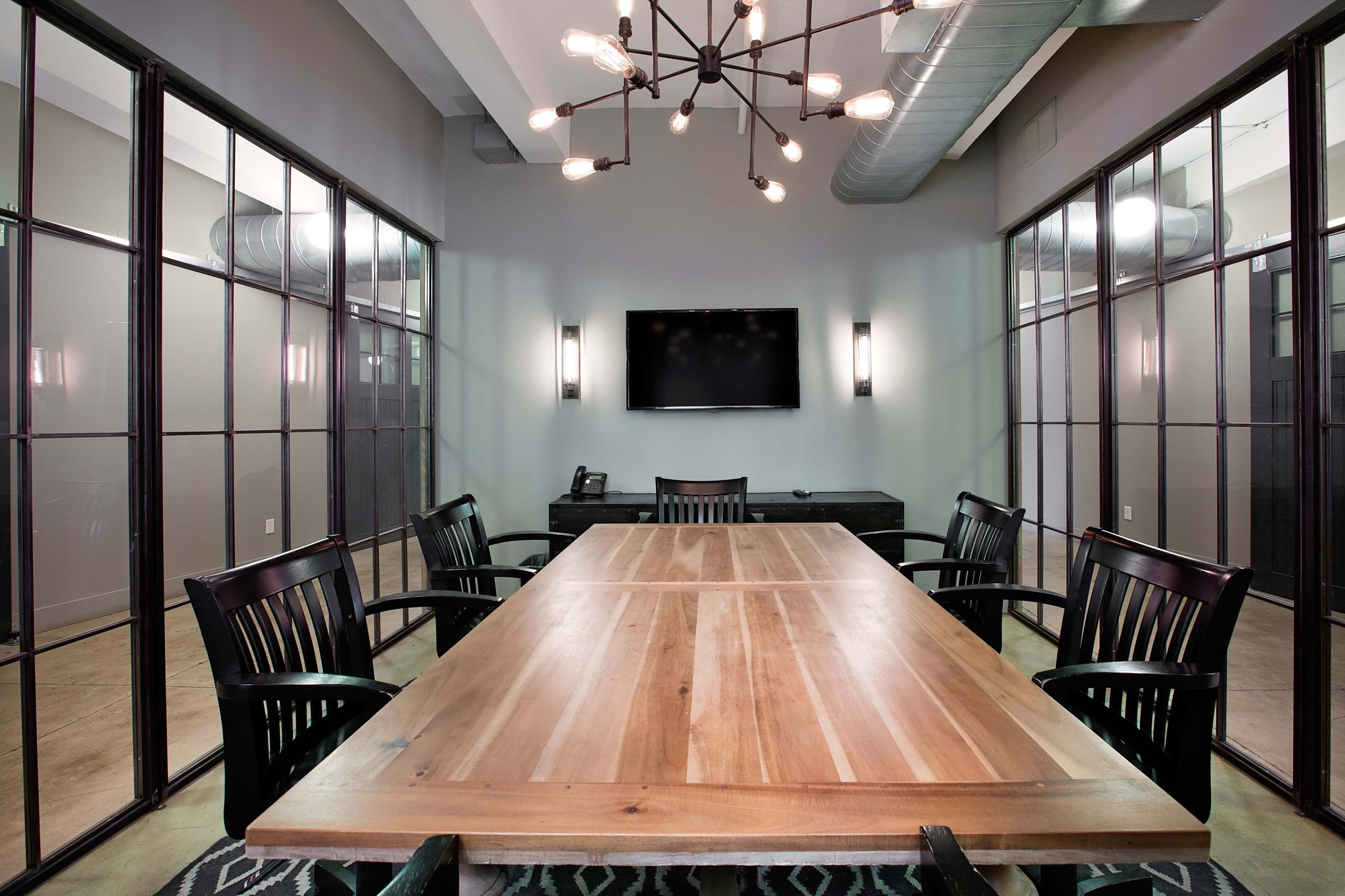 The image depicts a modern conference room featuring a large wooden table surrounded by black chairs, with glass-paneled walls and a wall-mounted TV.