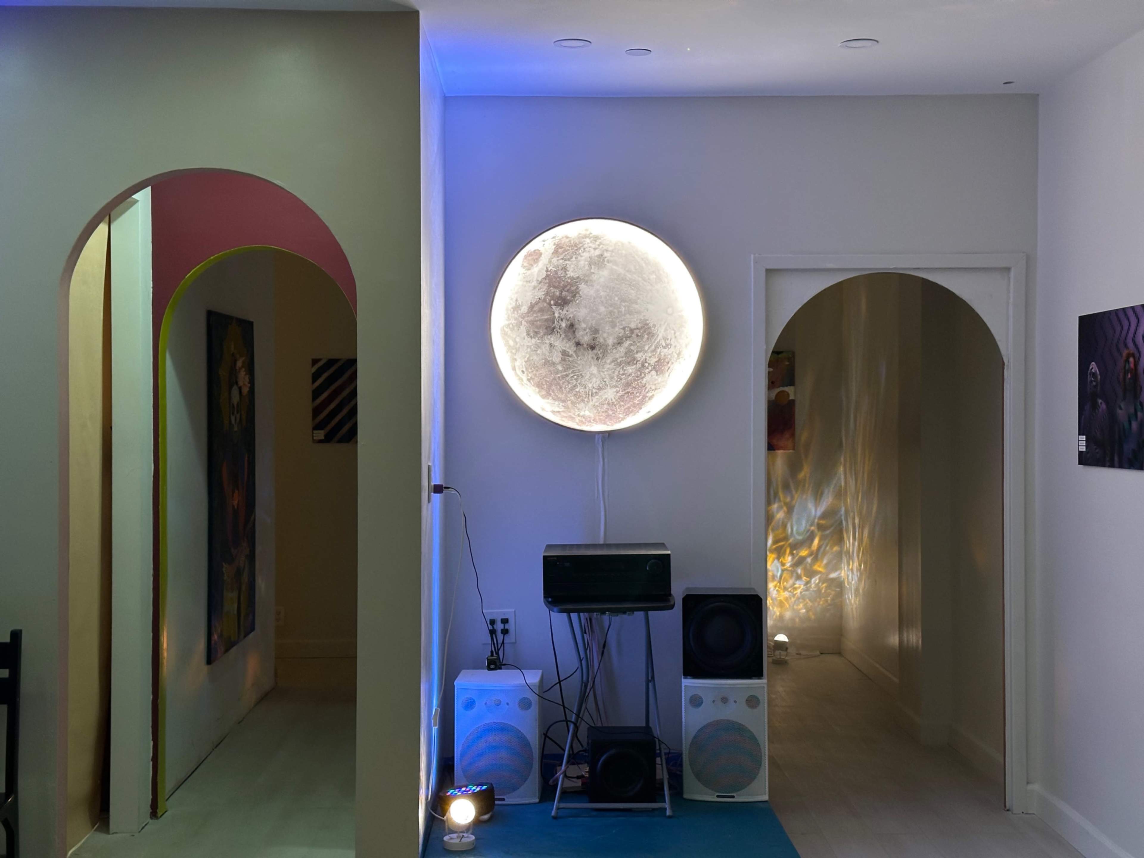 The image shows a modern interior with two arches leading to separate hallways, illuminated by a large circular moon lamp on the wall and sound equipment placed on the floor.