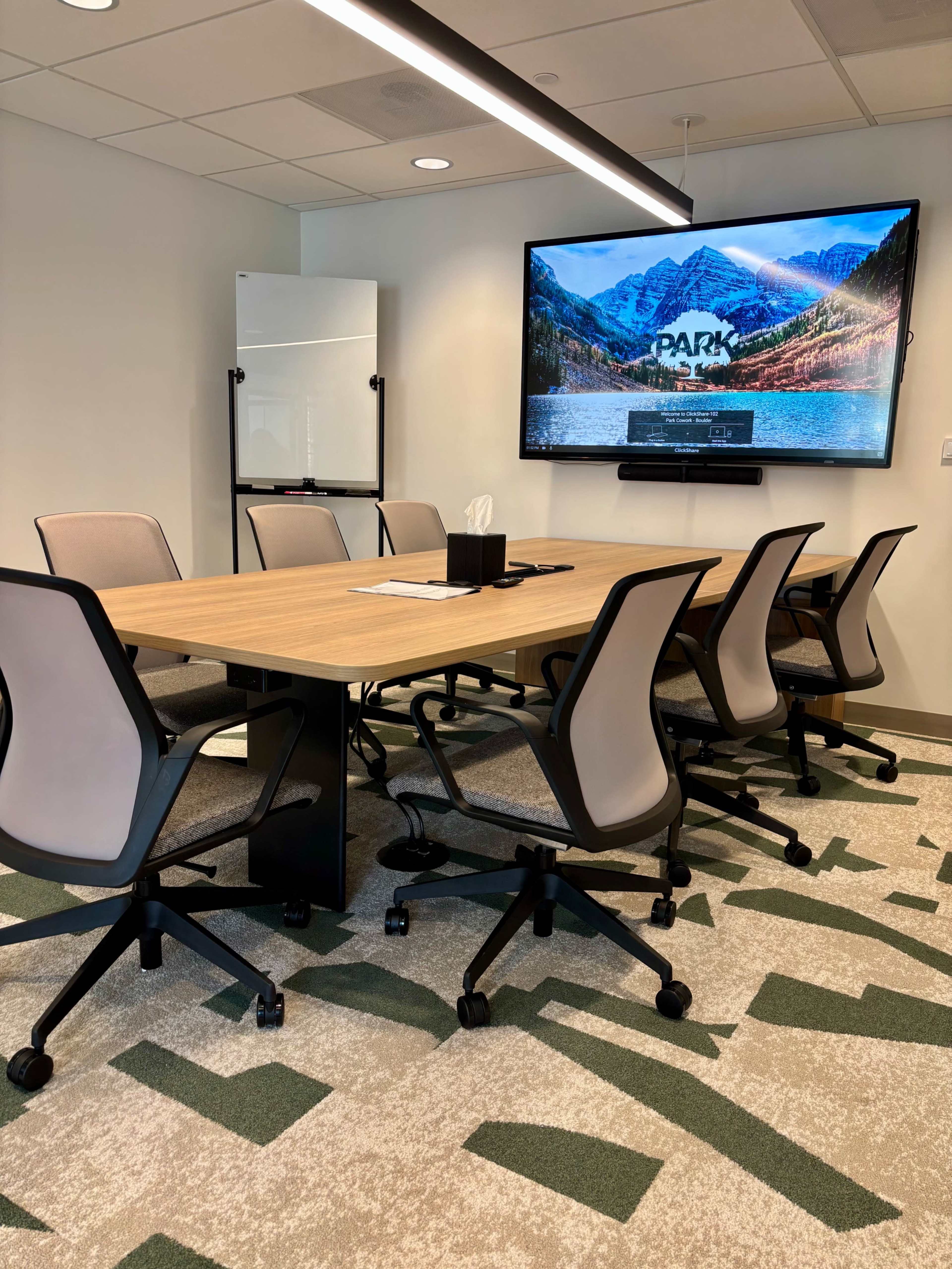 A modern conference room features a wooden table surrounded by ergonomic chairs, a large screen on the wall, and a whiteboard.