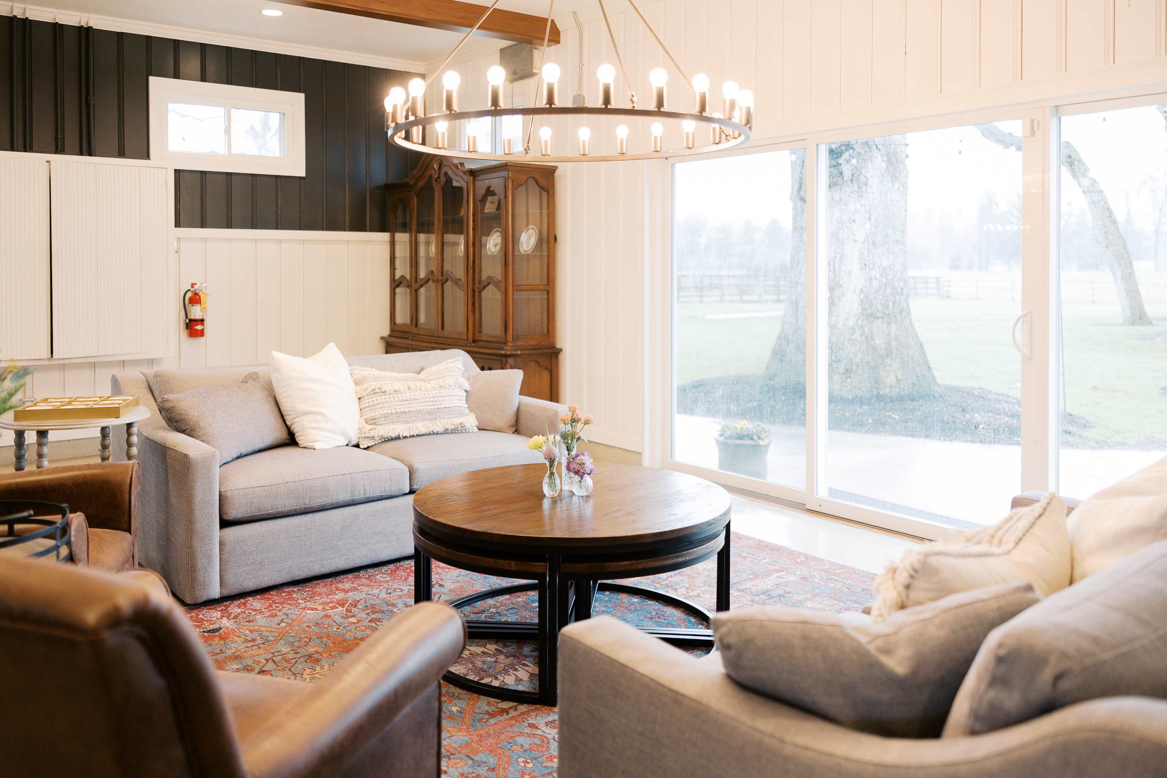 The image shows a cozy living area with two gray sofas, a round wooden coffee table, and a chandelier, set against a backdrop of large windows overlooking a grassy outdoor area.