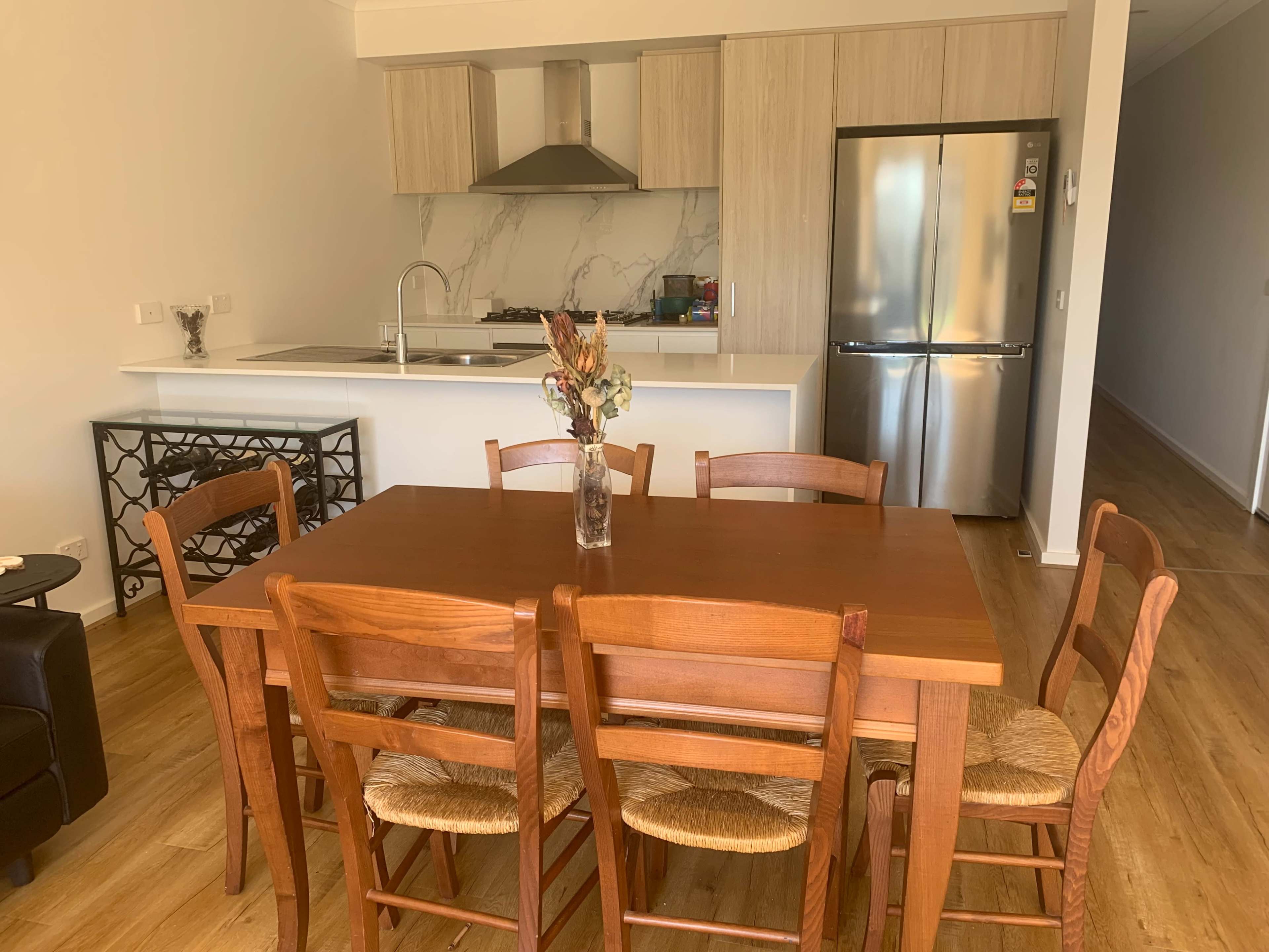 A wooden dining table with six chairs is set in a modern kitchen featuring a marble backsplash and stainless steel appliances.