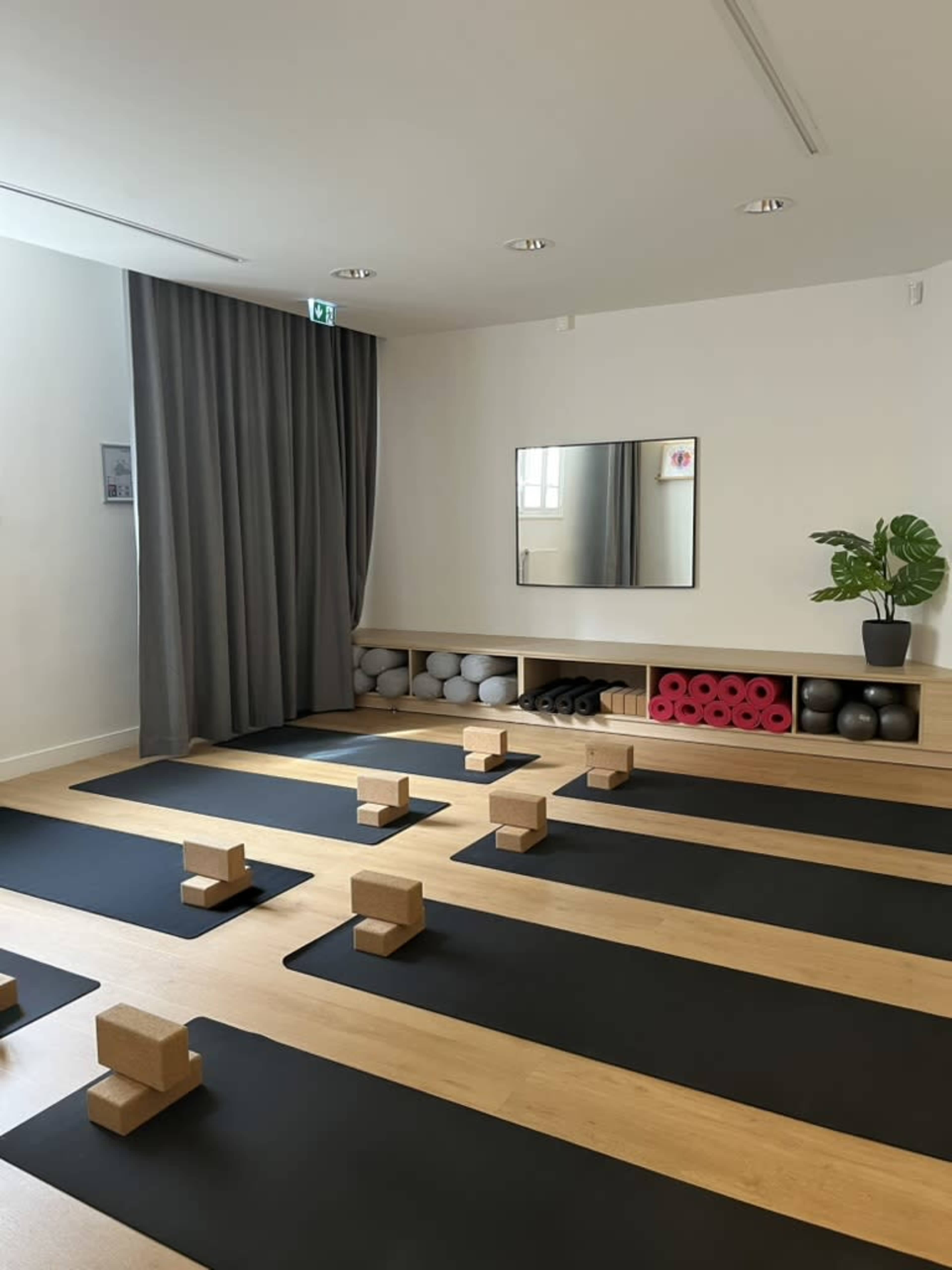 The image shows a well-lit yoga studio featuring black mats arranged neatly on the floor, with yoga blocks placed on each mat and exercise accessories stored on shelves.