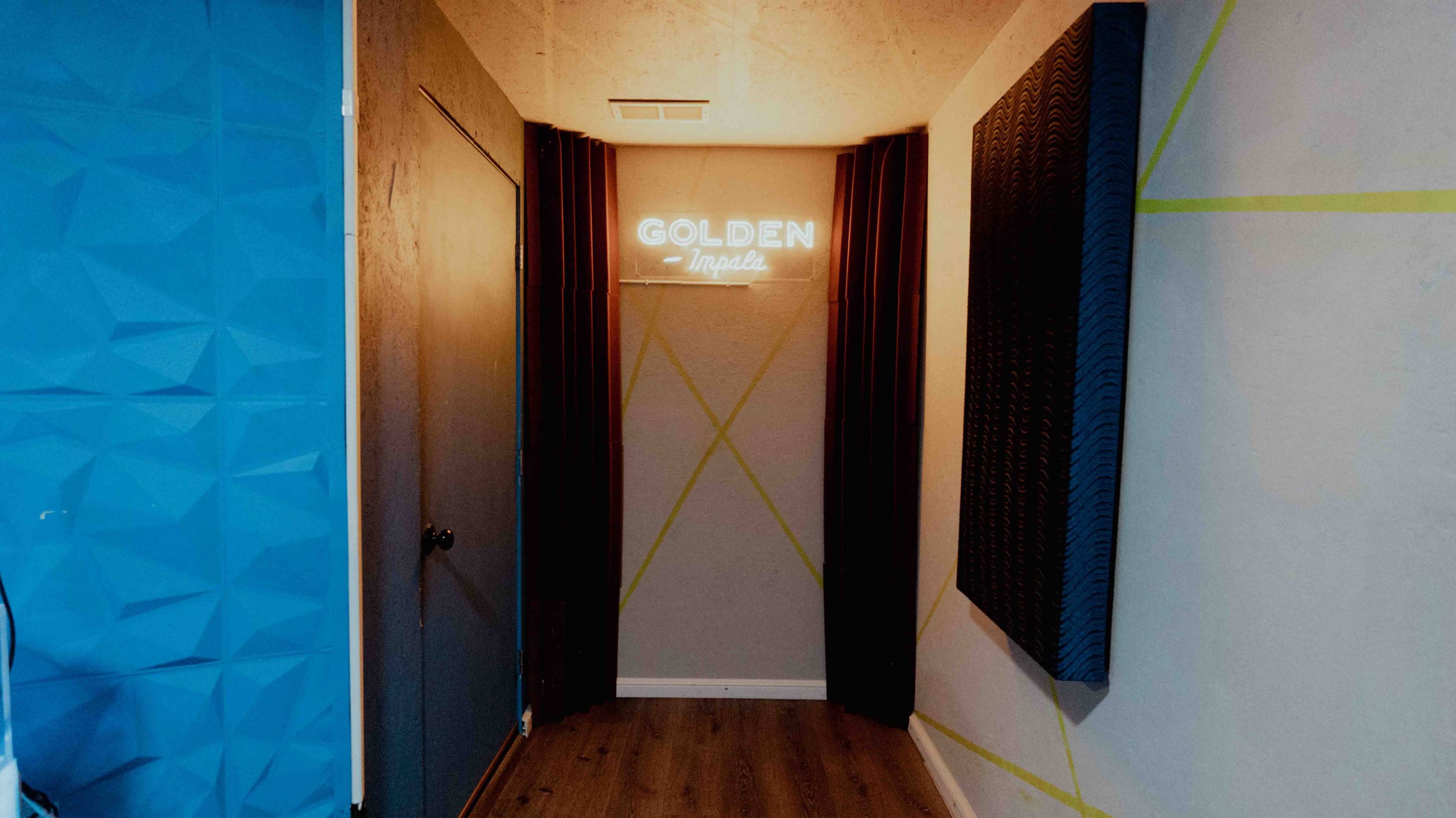 The image shows a narrow hallway with a neon sign reading "GOLDEN Impala," flanked by curtains and textured walls, leading to a closed door.
