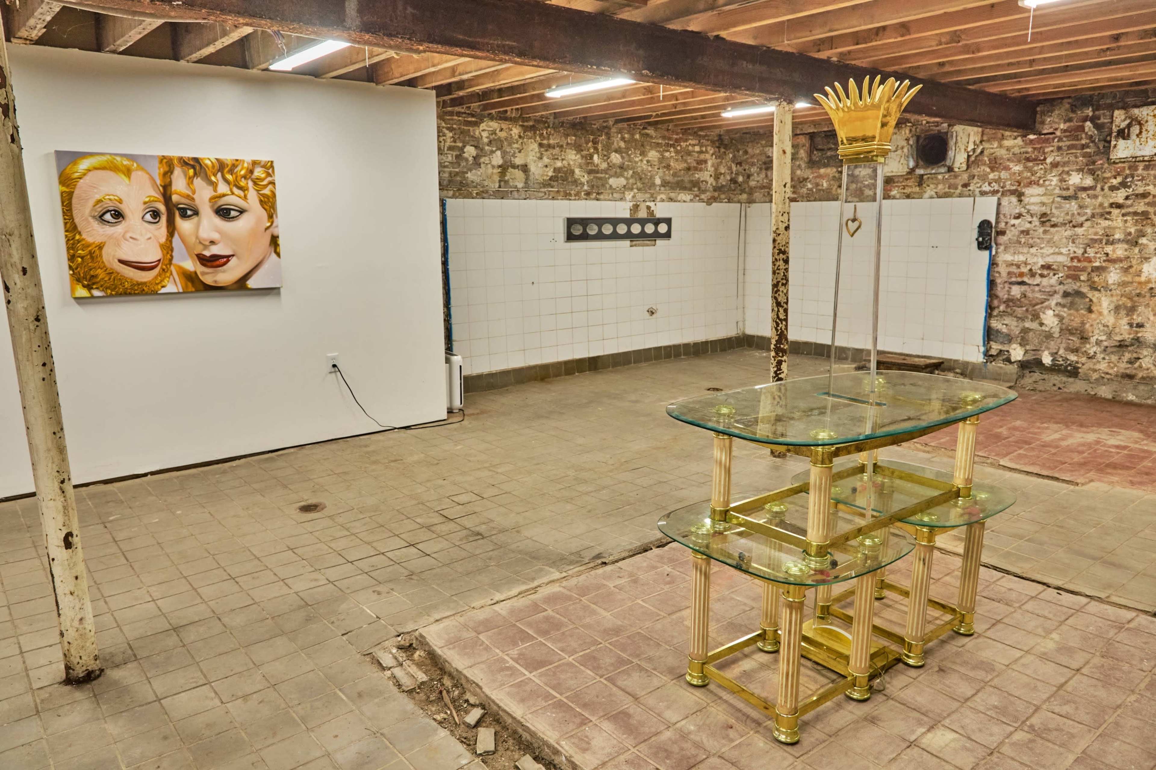 The image shows an empty basement space featuring a glass display table, a wall with circular objects, and a striking painting of two faces on the adjacent wall.