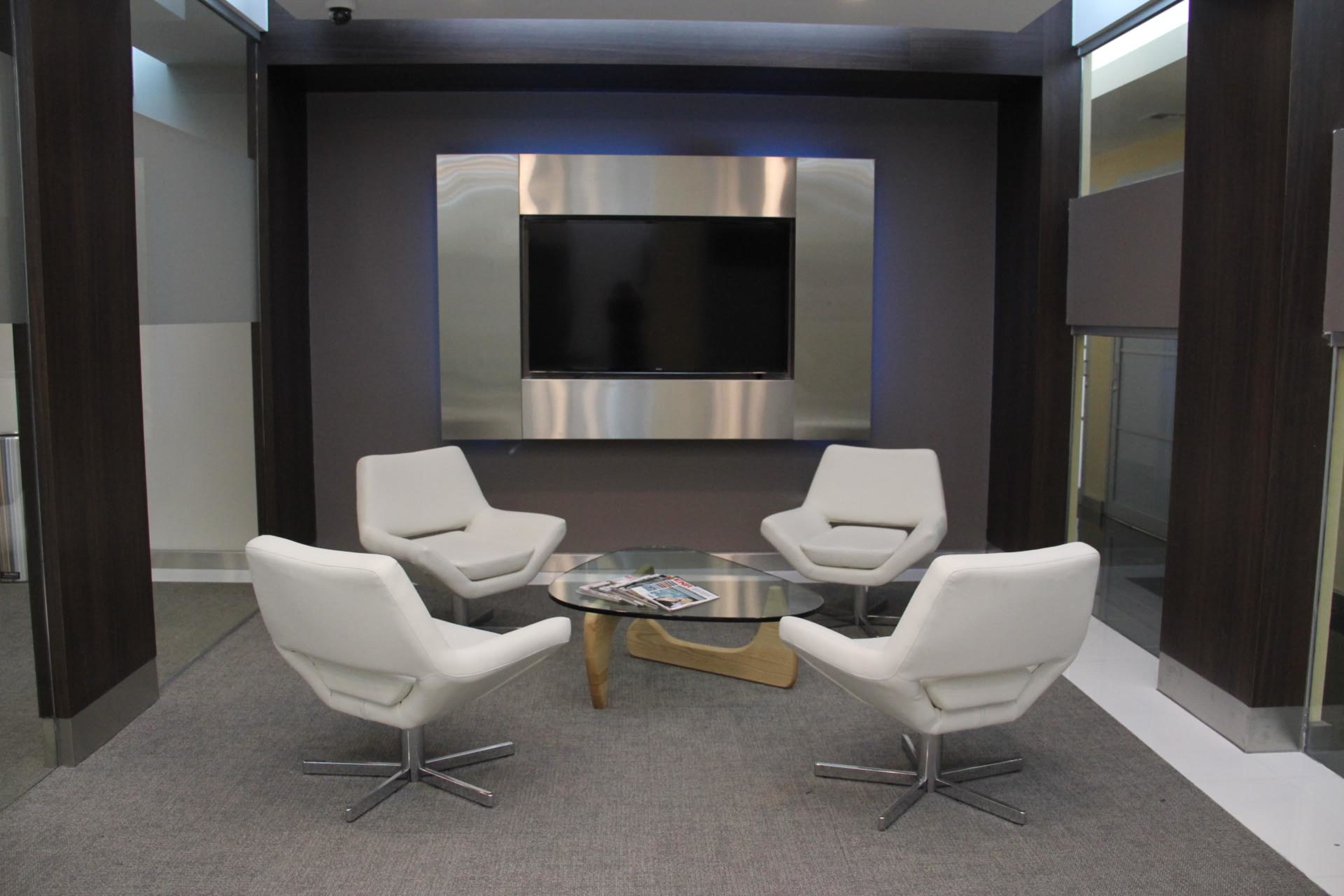A modern seating area features four white chairs arranged around a glass coffee table, set against a backdrop with a mounted television.