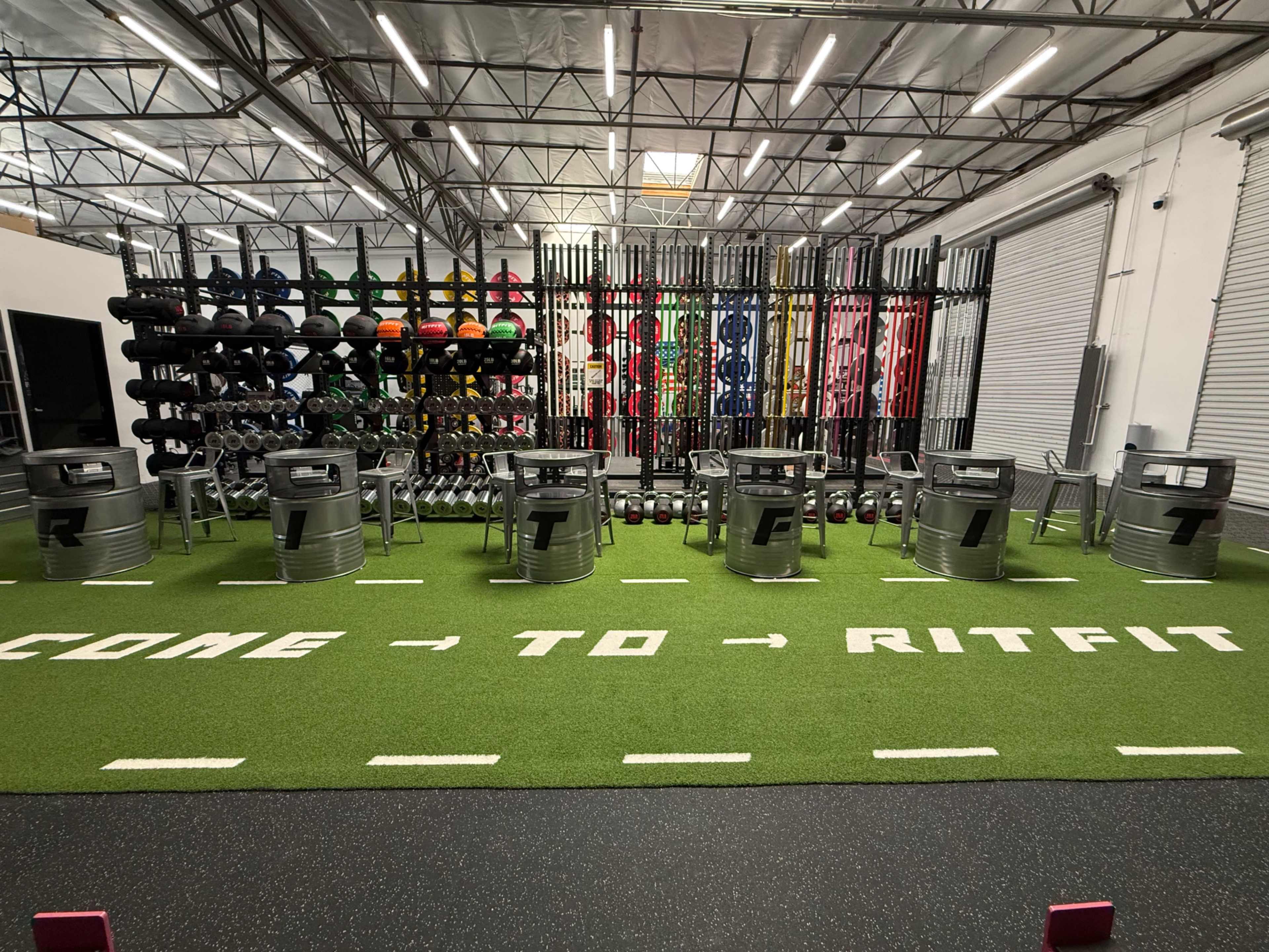 The image shows a gym interior with a green turf floor, metal containers arranged in a circle, and a wall of colorful weights and dumbbells in the background.