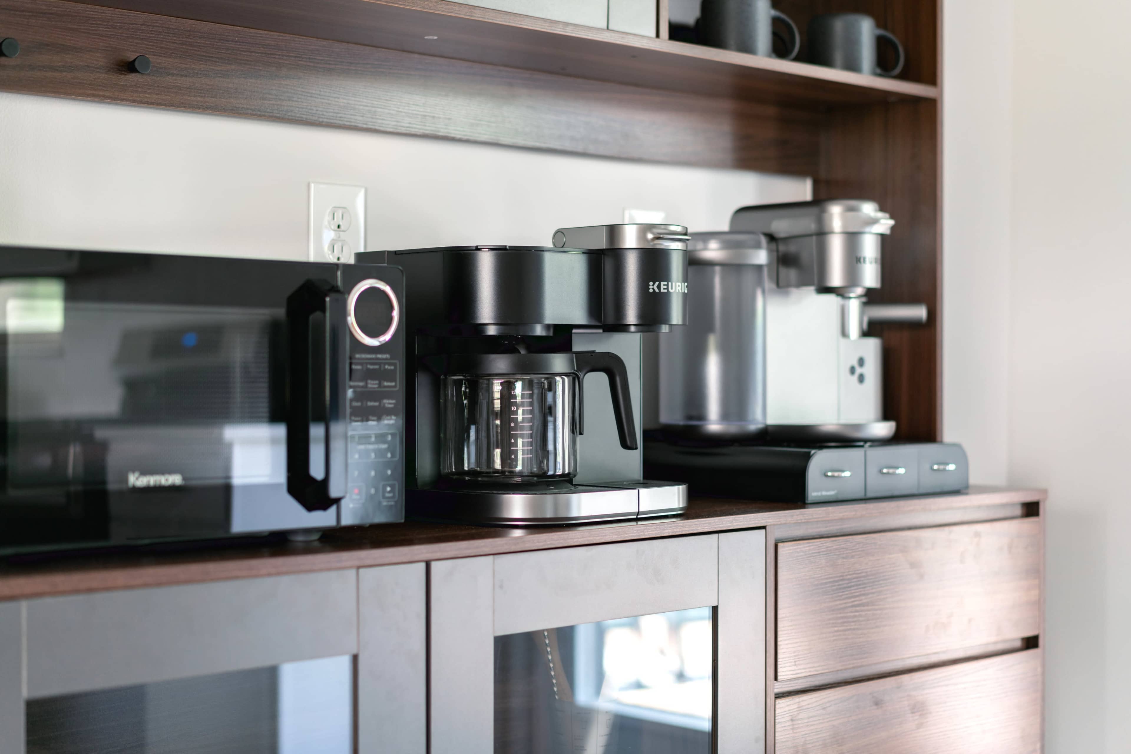 A kitchen countertop with a coffee maker, a microwave, and a single-serve coffee machine arranged on wooden shelves.