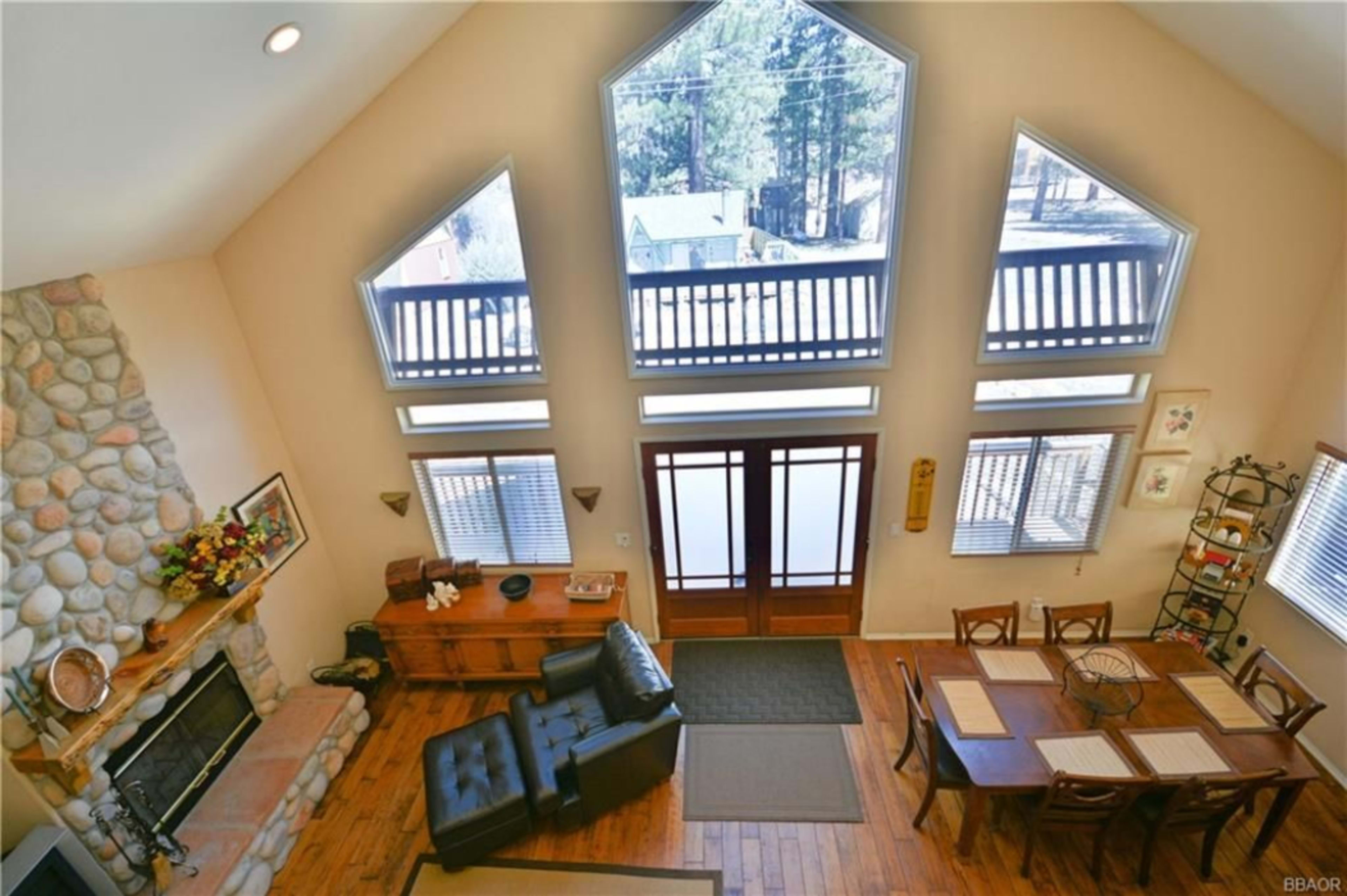 The image shows a spacious living area with a stone fireplace, large triangular windows, and a wooden dining table, all illuminated by natural light.