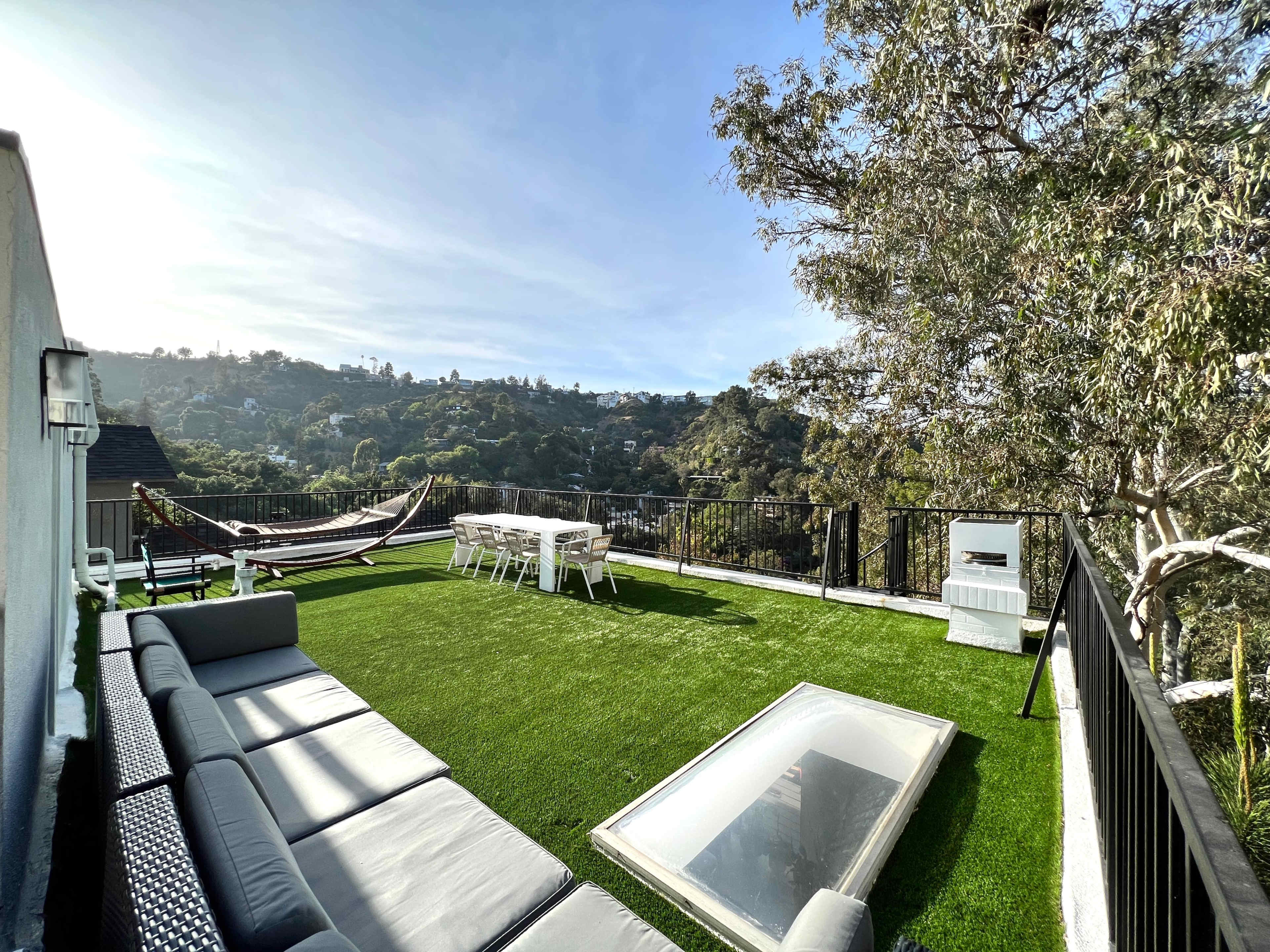 The image shows a rooftop patio with a green lawn, a hammock, a dining table, and a sectional sofa overlooking a hilly landscape.