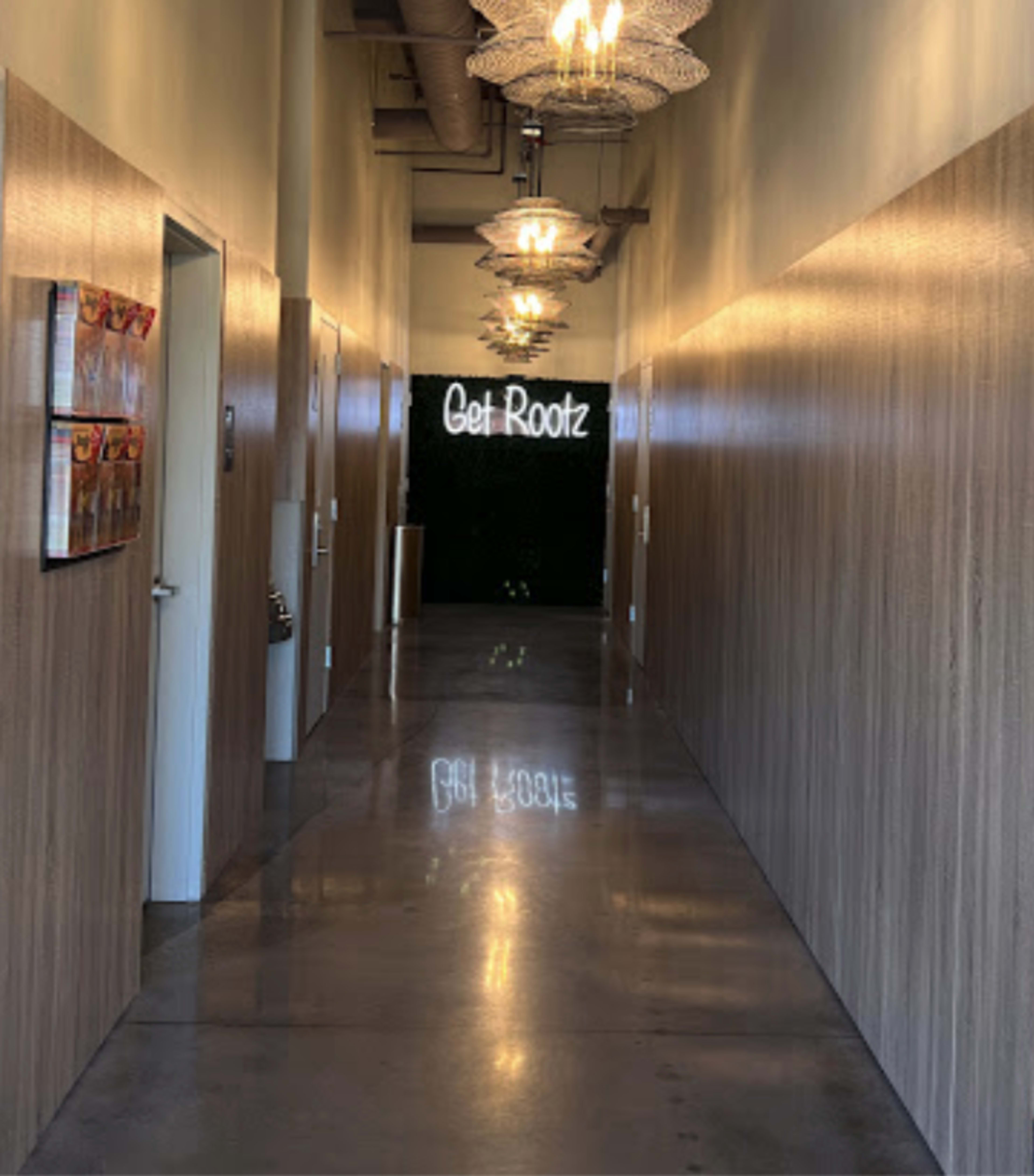 A long hallway features wooden paneling, overhead chandeliers, and a neon sign reading "Get Rootz" at the far end.