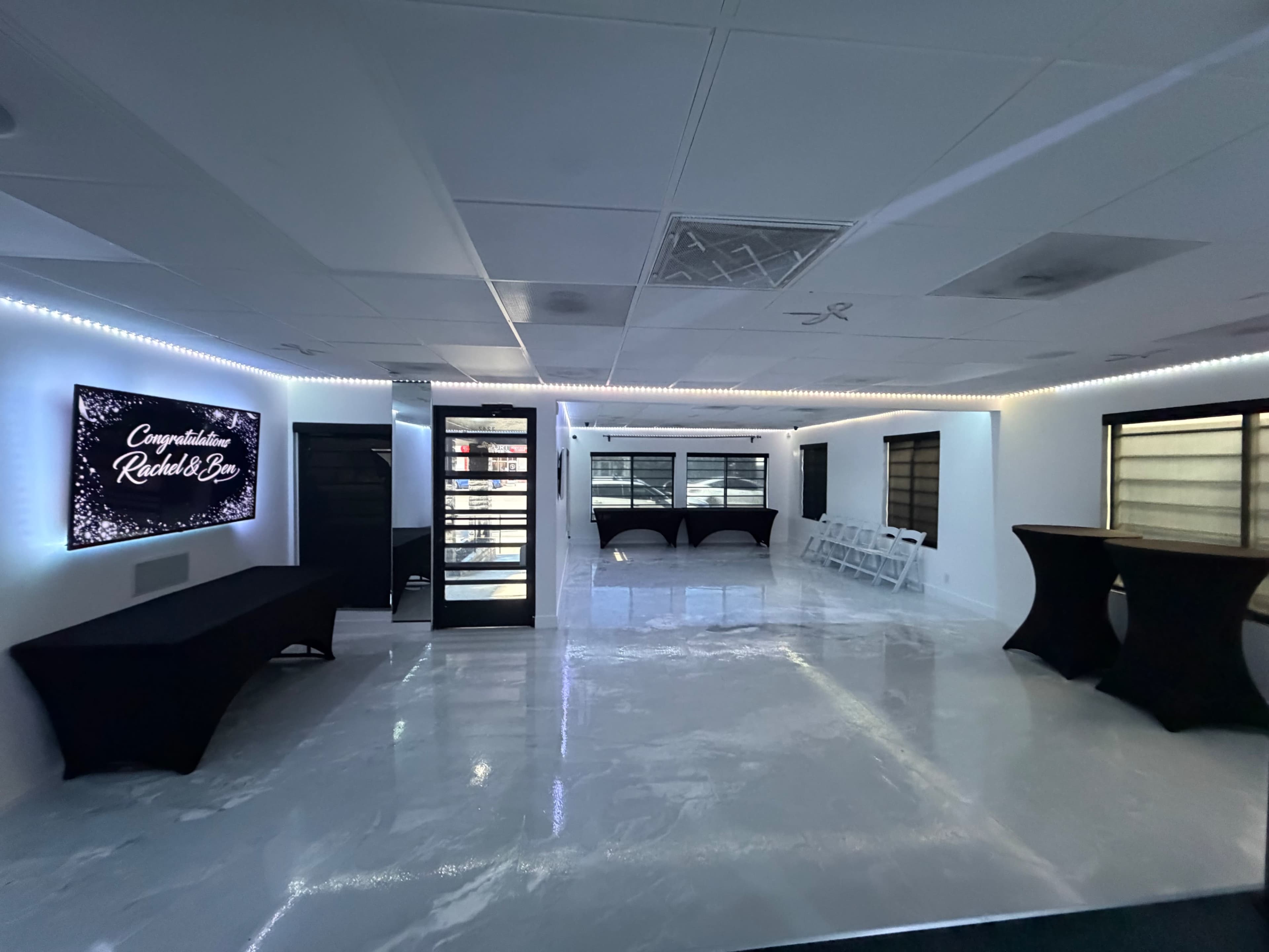 The image shows an empty, brightly lit event space with glossy white floors, black tables, and a digital screen displaying a congratulatory message.