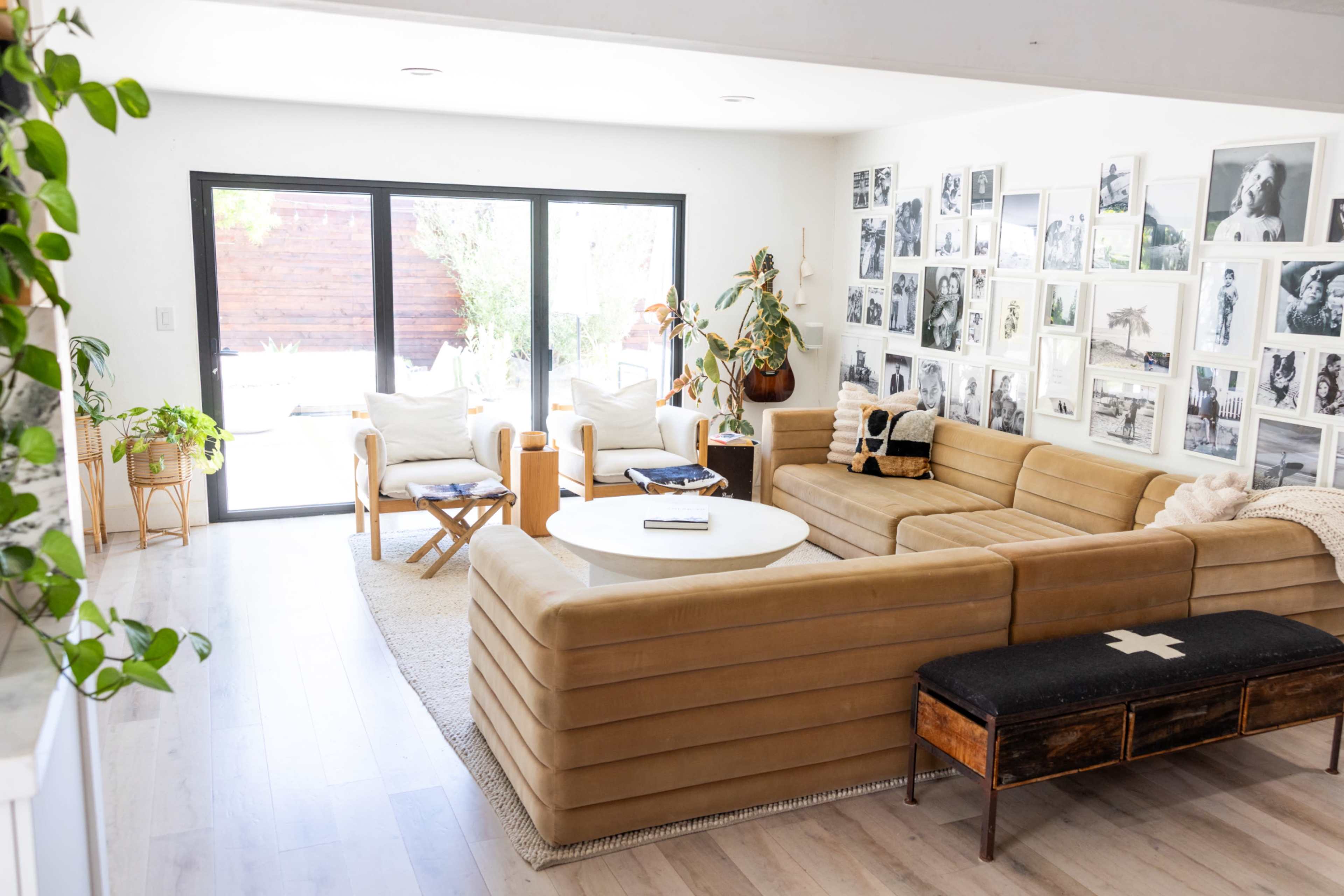 The living room features a tan sectional sofa, a round coffee table, and a wall adorned with framed photographs, with large sliding doors opening to an outdoor area.