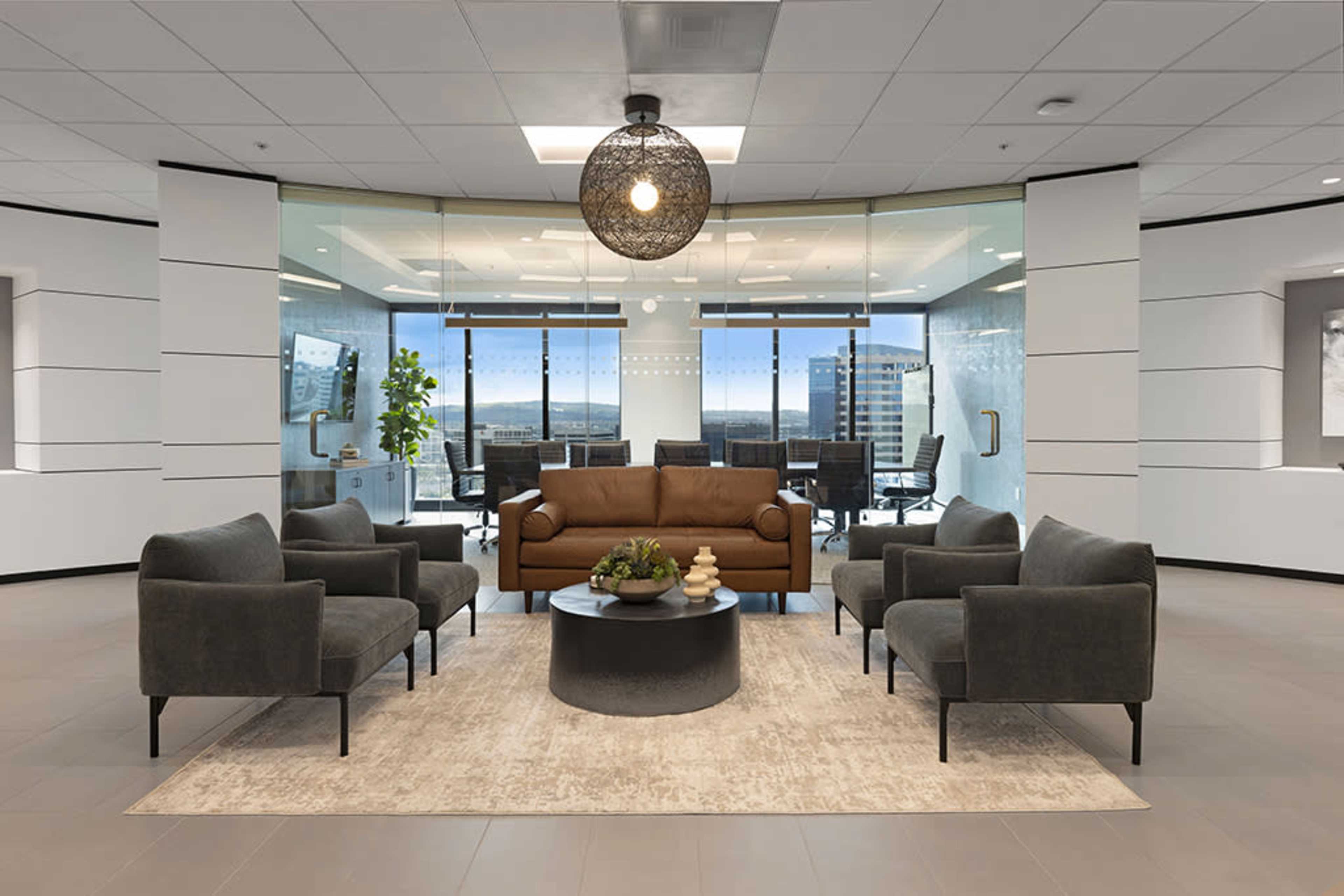 A modern office reception area features a brown leather sofa, four gray armchairs, a round coffee table, and large glass windows overlooking a cityscape.