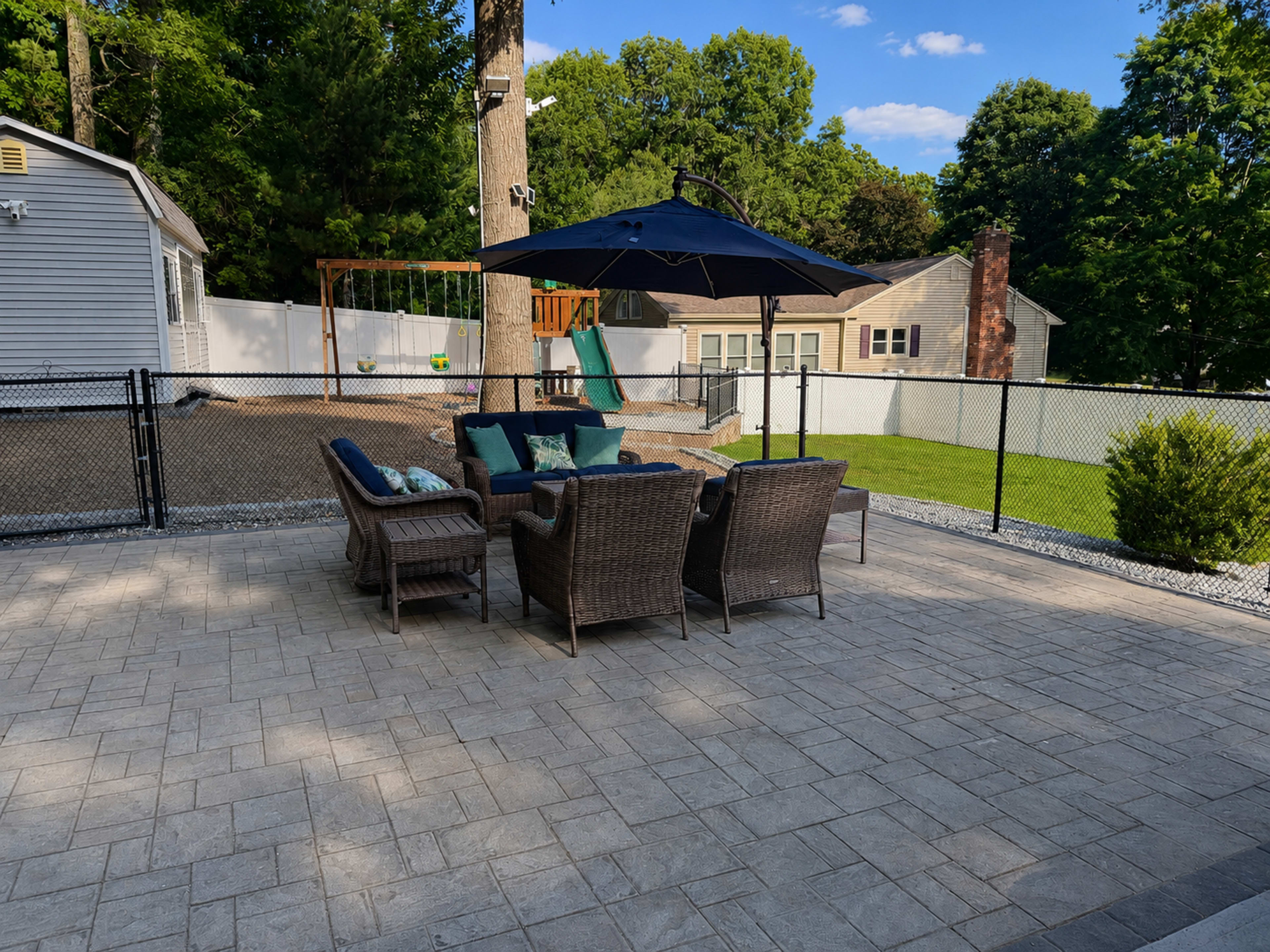 A backyard patio features a set of wicker chairs and a table under a large umbrella, with a swing set visible in the background.
