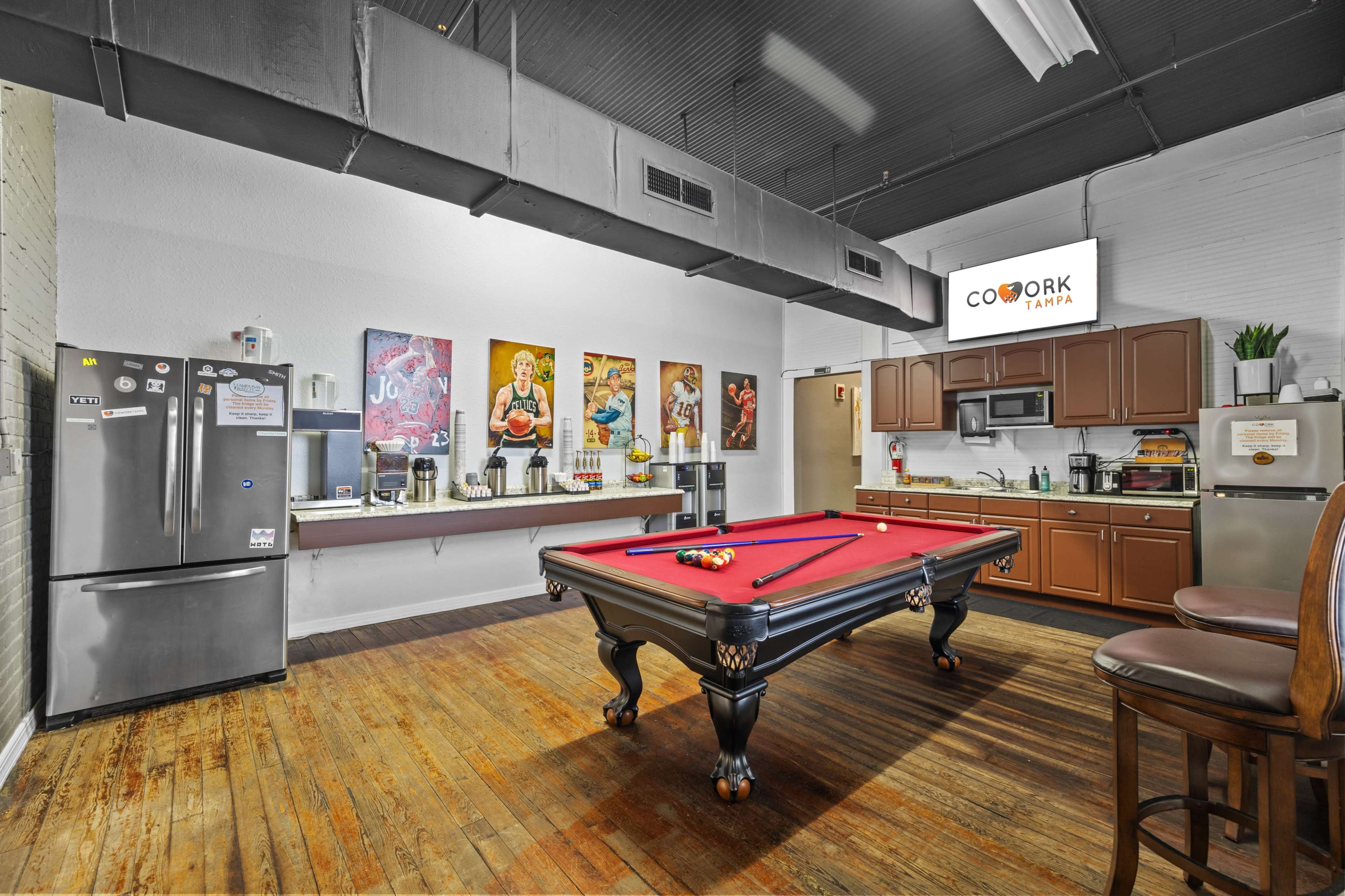 The image shows a kitchenette area with a pool table in the center, surrounded by wooden flooring and decorated with several framed artworks on the walls.