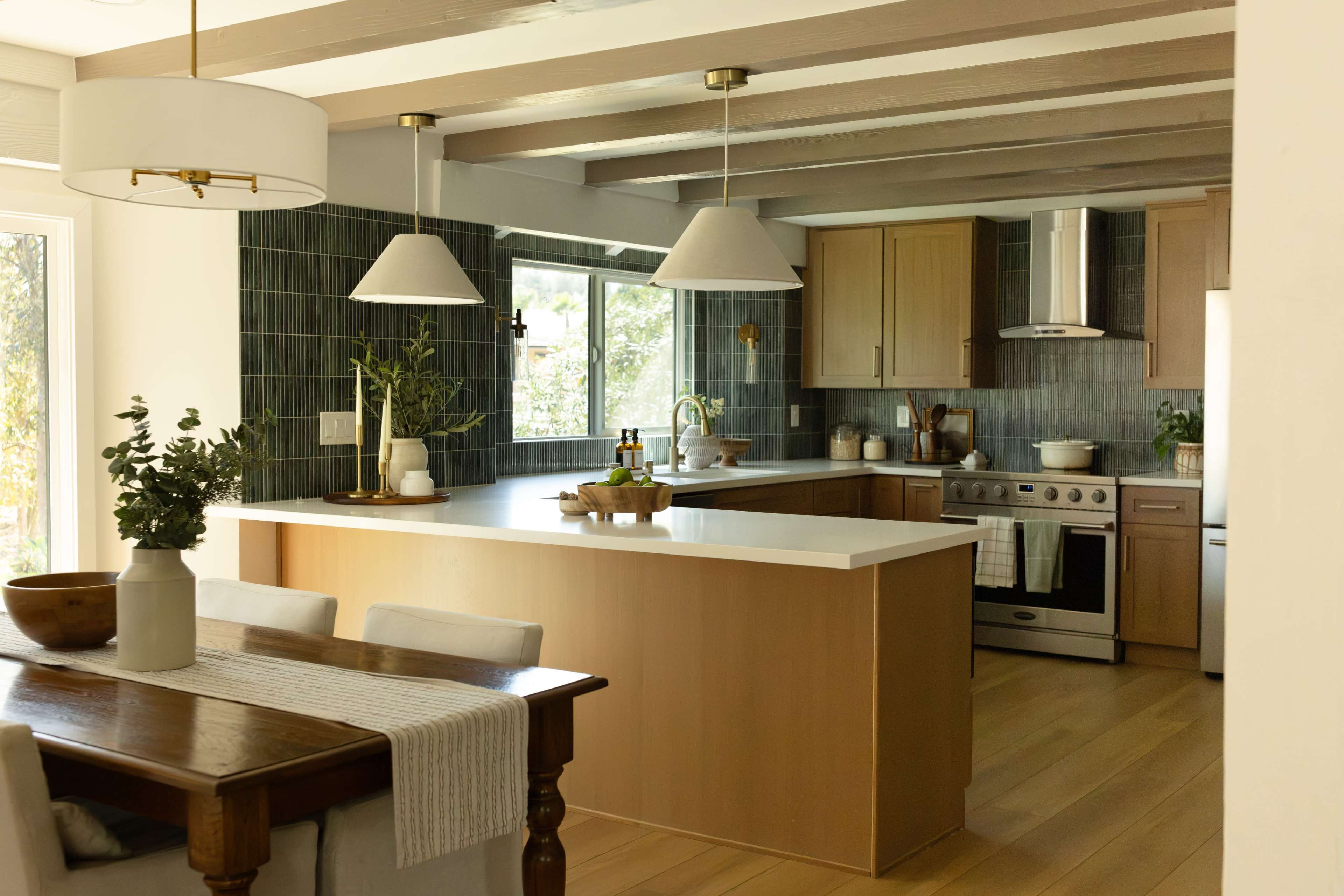 The image shows a modern kitchen with a central island, white countertops, wooden cabinetry, and grey tiled walls, featuring a dining area with a wooden table.
