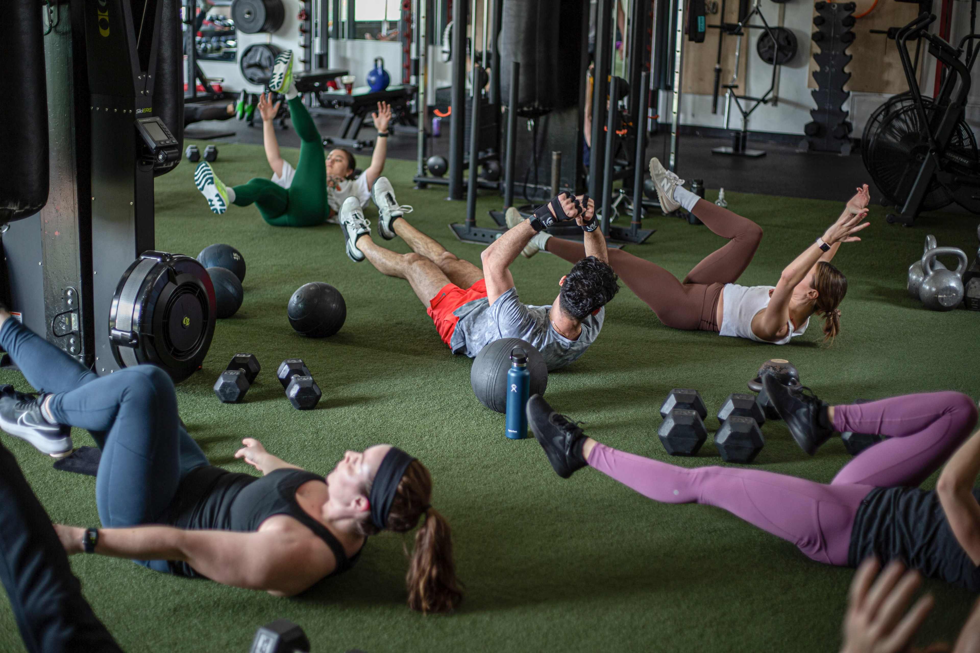 A group of people perform various exercises on a gym floor, utilizing weights and exercise balls.